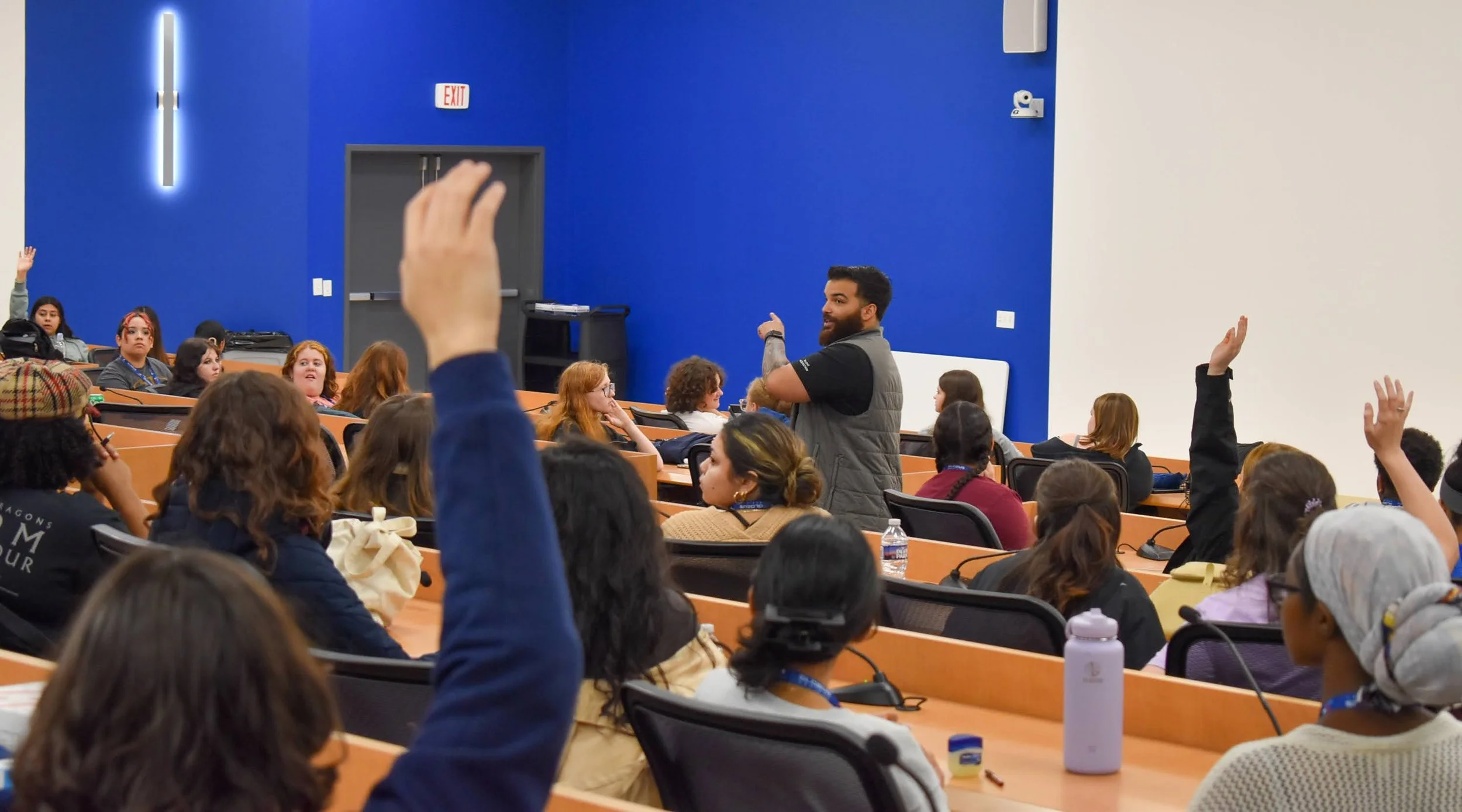 A man standing at the front of a classroom or conference room, engaging with students or attendees who are seated and raising their hands, with some taking notes. The room has a blue and white wall background, and attendees are diverse, including women and children.