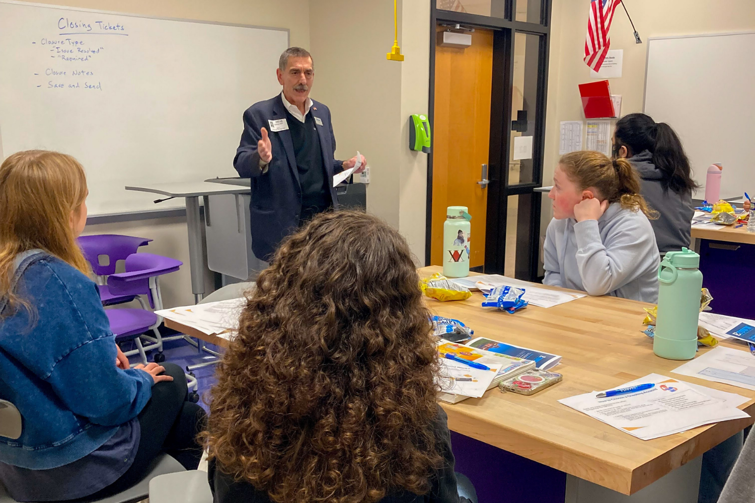 A man in a suit is standing at the front of a classroom, speaking to a group of students seated at a table, with a whiteboard behind him. The whiteboard has notes about closing tickets, and the students have snacks, water bottles, and papers on the table.