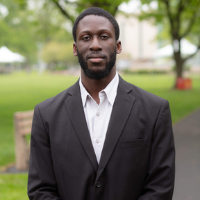 A man in a black suit and white shirt standing outdoors in a park with trees and grass in the background.