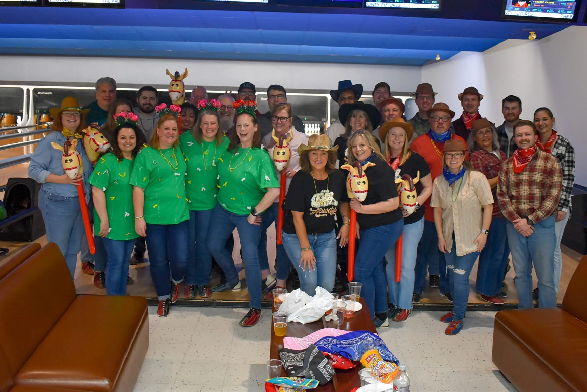Group of people dressed in cowboy and western themed costumes, some holding inflatable horse toys, inside a bowling alley.