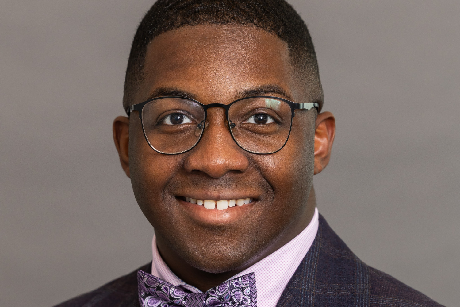 A smiling man with glasses, wearing a checkered suit, pink shirt, and purple patterned bow tie, against a plain background.
