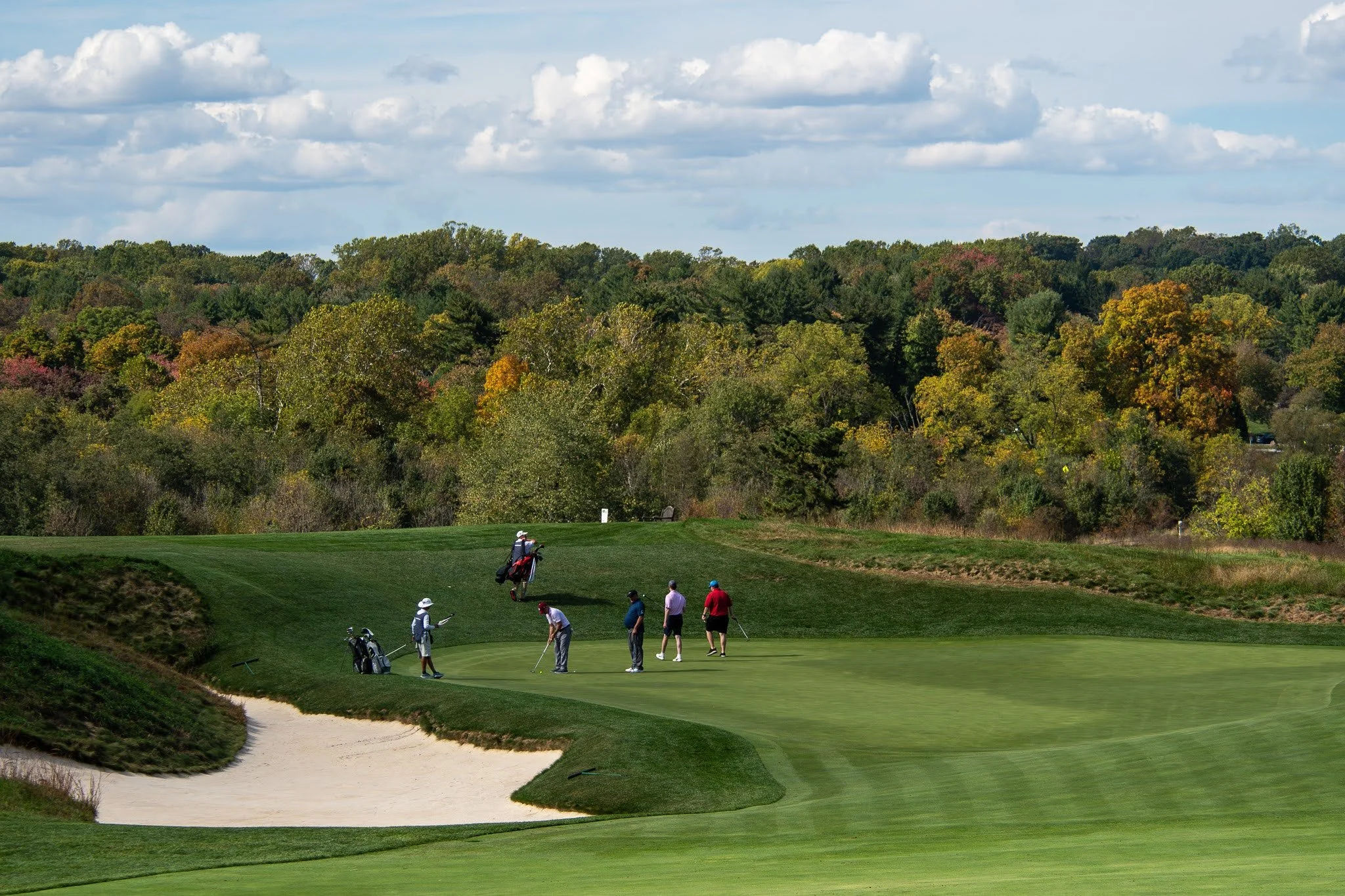 Golfers on a putting green preparing to putt near a sand trap on a golf course, with trees showing fall colors and a partly cloudy sky in the background.