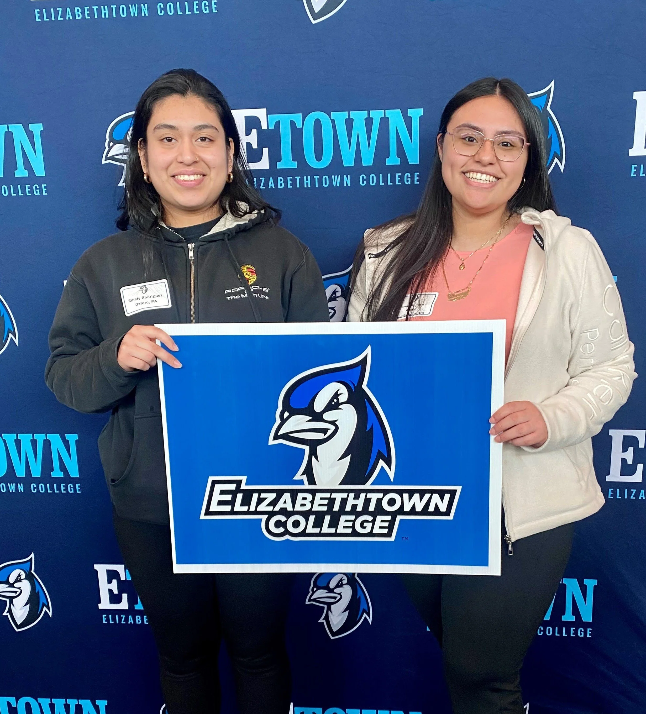 Two smiling women holding a large Elizabethown College sign at a college event, standing in front of a blue backdrop with the Elizabethown College logo. One woman wears glasses and a beige hoodie, the other wears a black jacket.