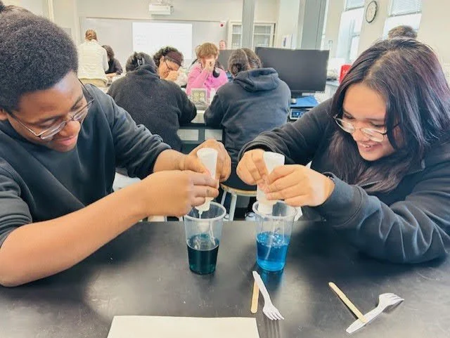 Two students in a classroom pouring blue and black liquids from white cups into clear glasses, smiling and working together.