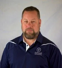 A man with a beard and short hair, wearing a navy blue polo shirt with white and blue accents. The shirt has a logo and text on the left side that says 'DeHewell'.