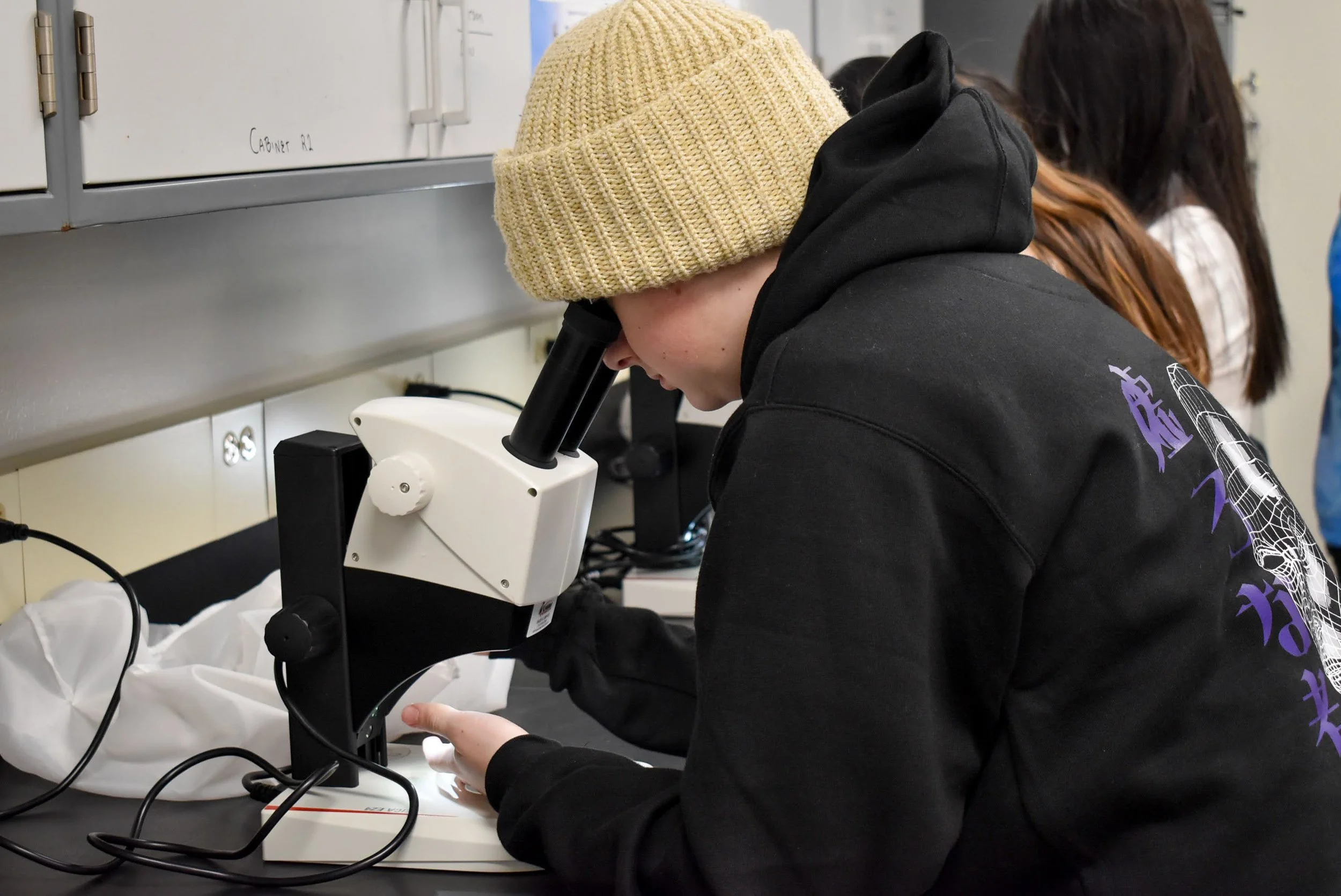 A person wearing a beige knit hat and black hoodie looking through a microscope in a laboratory.