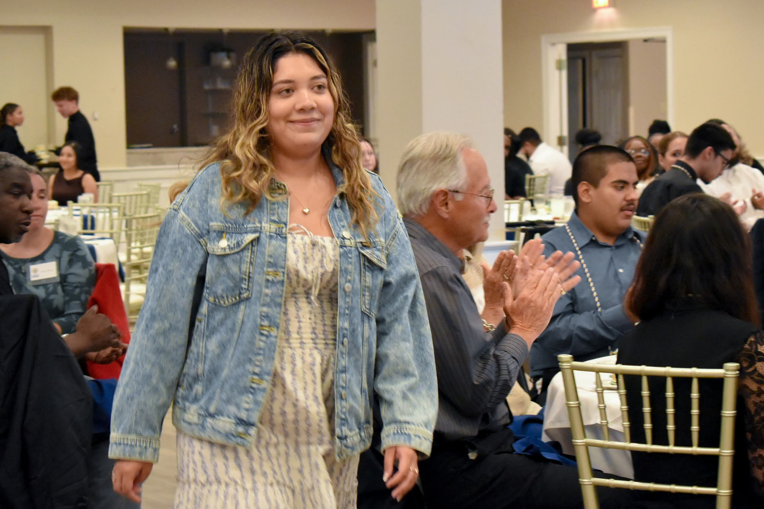 A young woman with wavy hair wearing a denim jacket and a dress, smiling and walking past seated guests at a formal event.