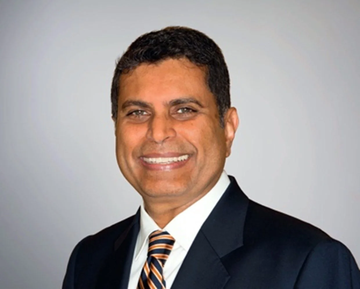 Professional headshot of a smiling man in a suit, white shirt, and striped tie, against a plain gray background.