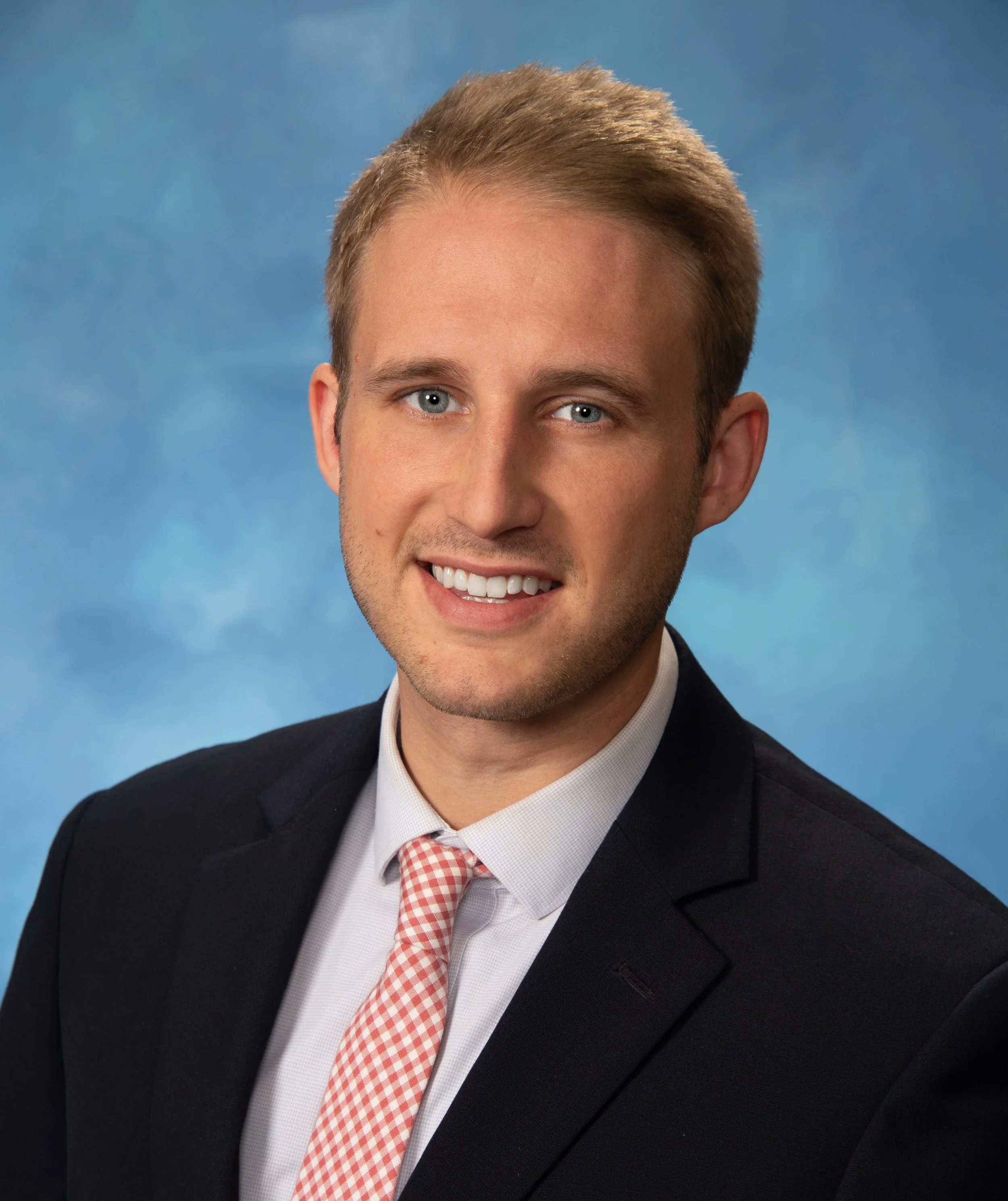 A young man with light brown hair, blue eyes, and light skin, wearing a black suit, white shirt, and a pink and white checkered tie, smiling against a blue background.