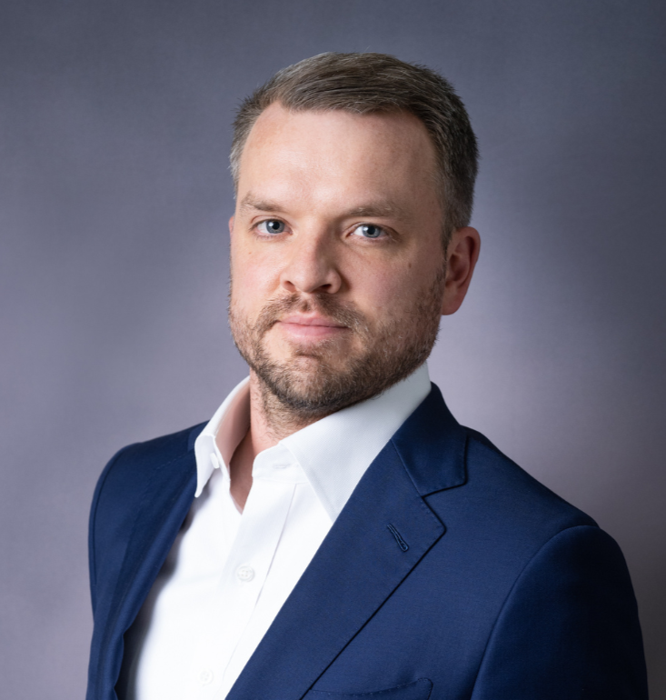 A man with short brown hair, a beard, dressed in a navy blazer and white shirt, posing against a gray background.
