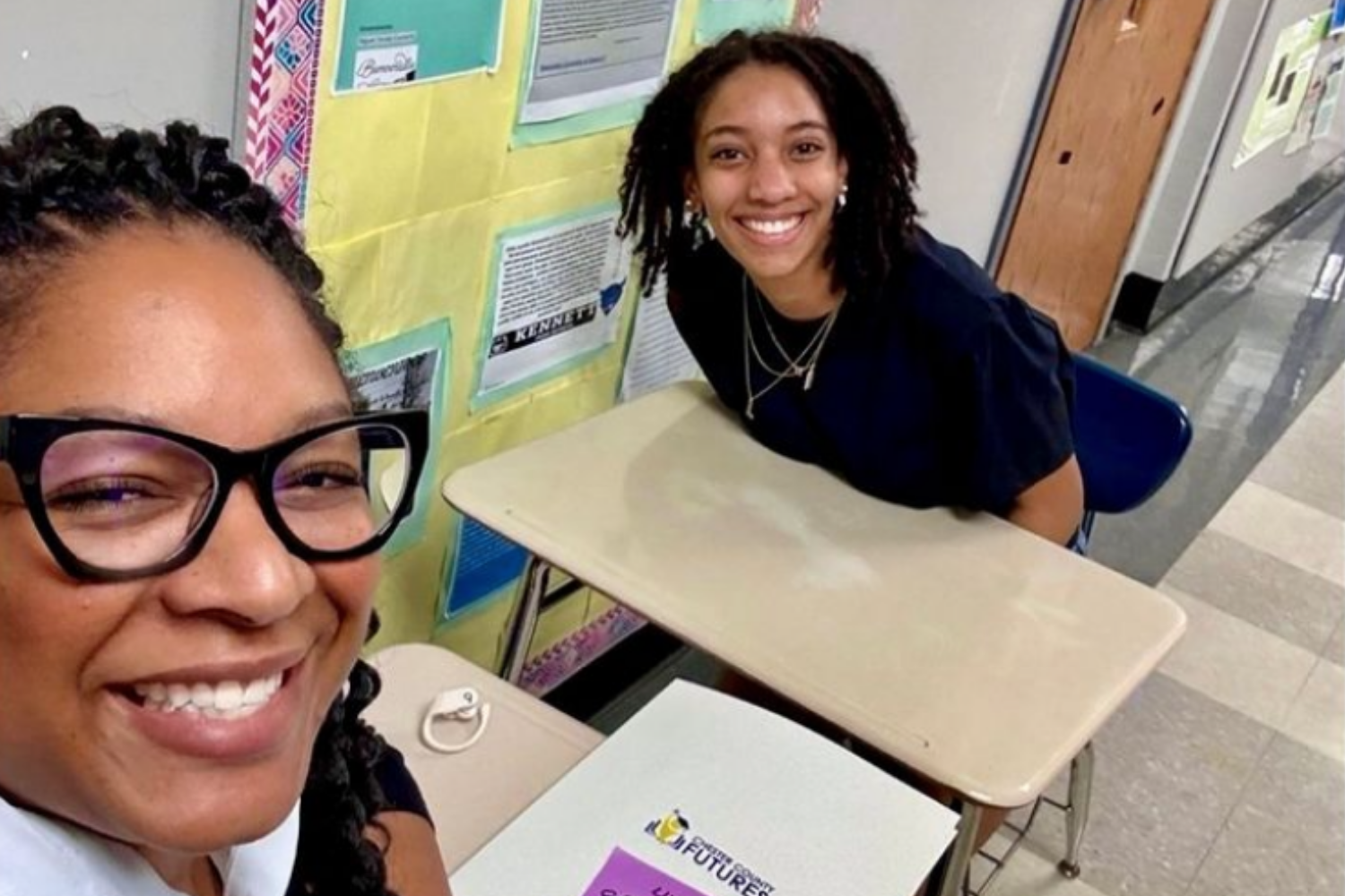 Two women smiling at a school desk in a hallway, with colorful bulletin boards and educational posters in the background.