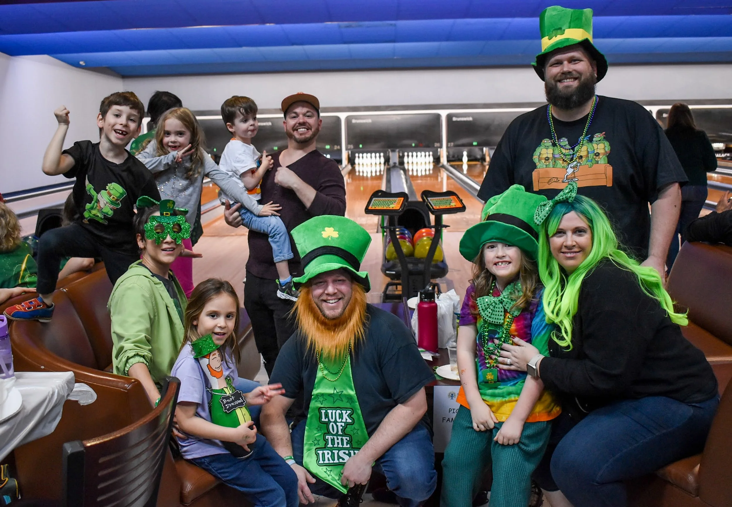 Group of children and adults celebrating St. Patrick's Day at a bowling alley, wearing green hats and accessories, with a bowling lane in the background.