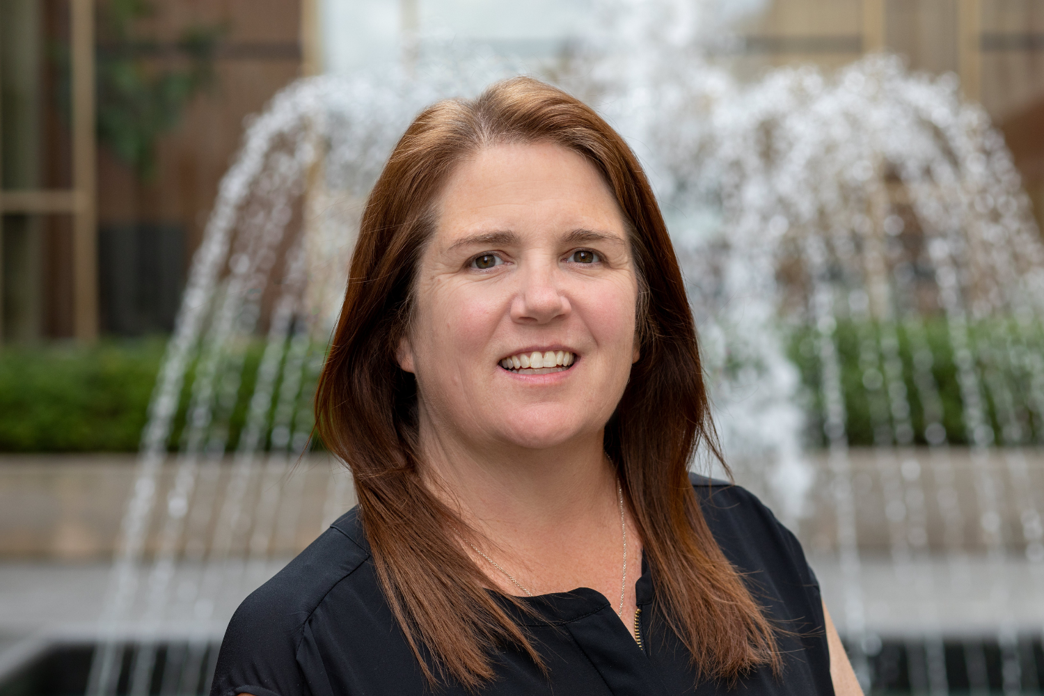A woman with shoulder-length brown hair and a black top, smiling in front of a water fountain with a blurred background of greenery and buildings.