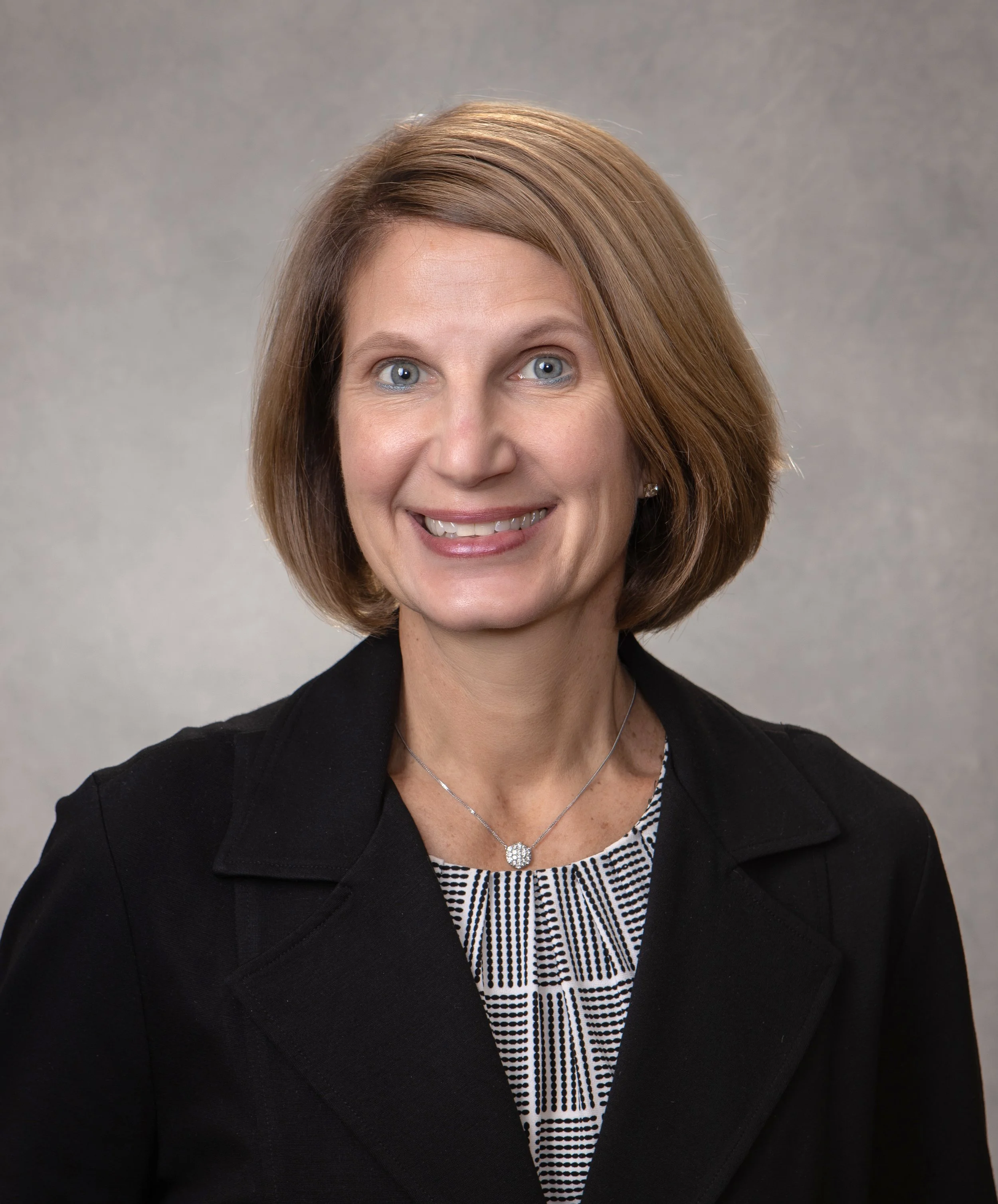 A portrait of a middle-aged woman with shoulder-length light brown hair, wearing a black blazer over a black and white patterned blouse, and a silver necklace, smiling at the camera against a plain gray background.