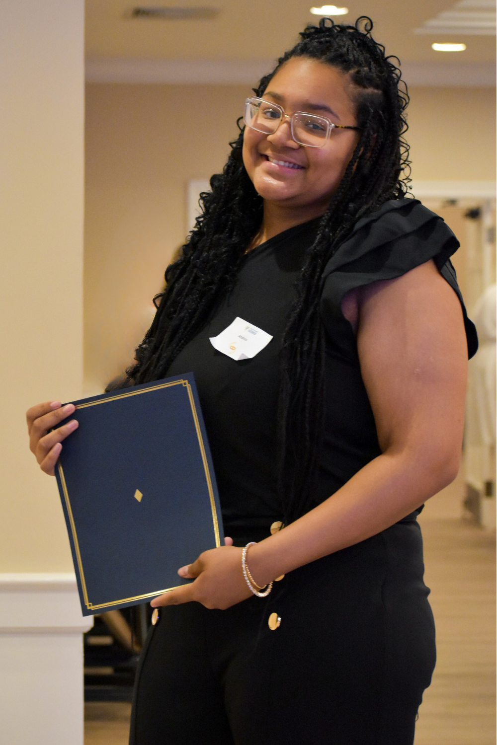 A smiling woman with glasses and long dreadlocks wearing a black top, holding a blue diploma cover, at an indoor event.