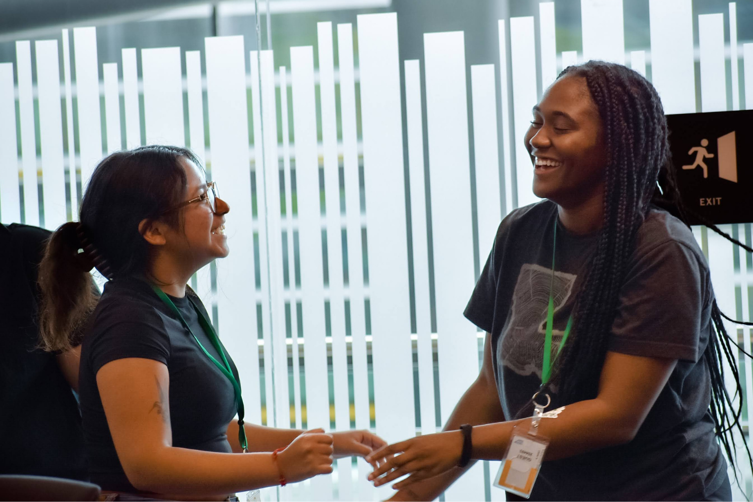 Two women smiling and holding hands at a conference or event, with a window and exit sign in the background.