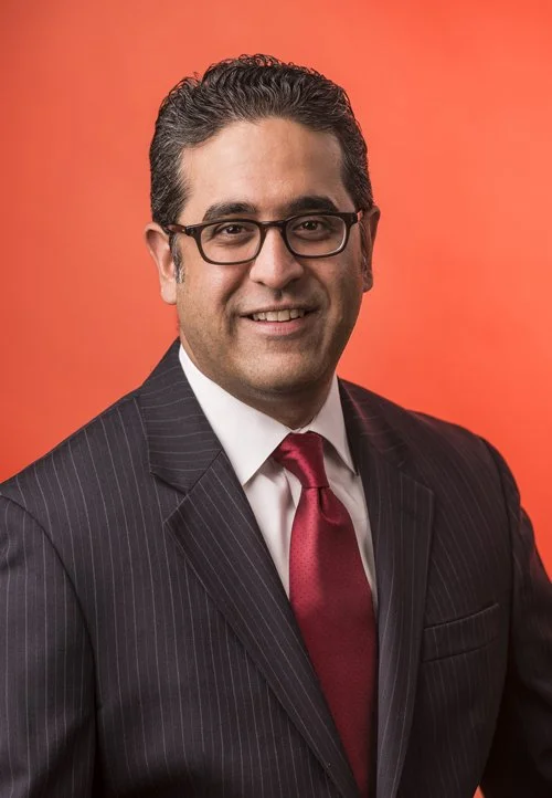 A man in a dark pinstriped suit, white shirt, red tie, and glasses, smiling against a red background.