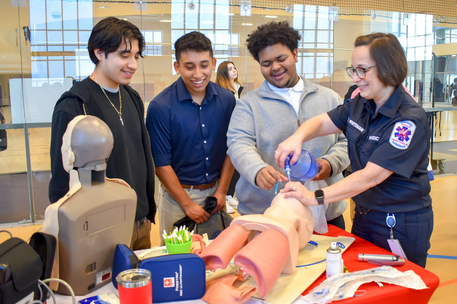 A medical professional demonstrating CPR on a mannequin to a group of four young men in a training room.