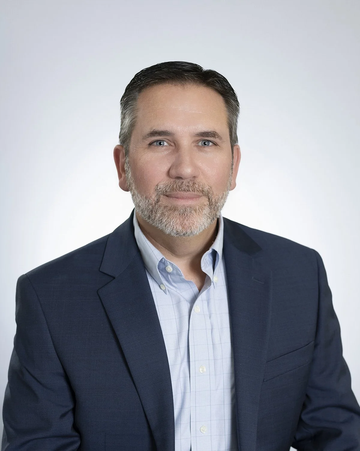 A middle-aged man with gray hair and beard wearing a navy suit and light blue button-up shirt, posing against a plain white background.