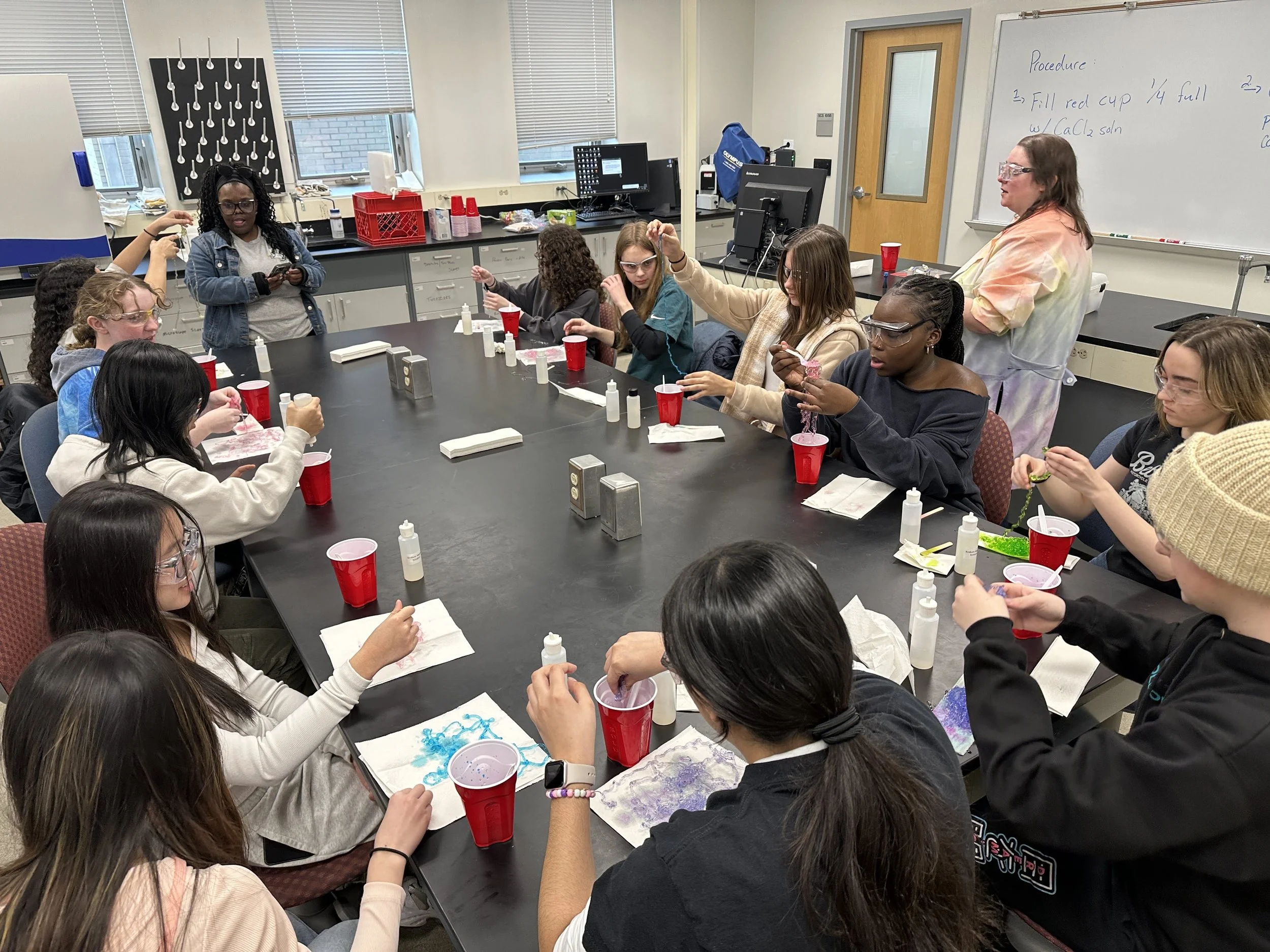 A classroom with students participating in a science activity. The students are seated around a large table, some wearing safety goggles, with bottles and supplies in front of them. A teacher is standing near a whiteboard with instructions written on it. There are windows in the background, and the room is equipped with computers and lab materials.