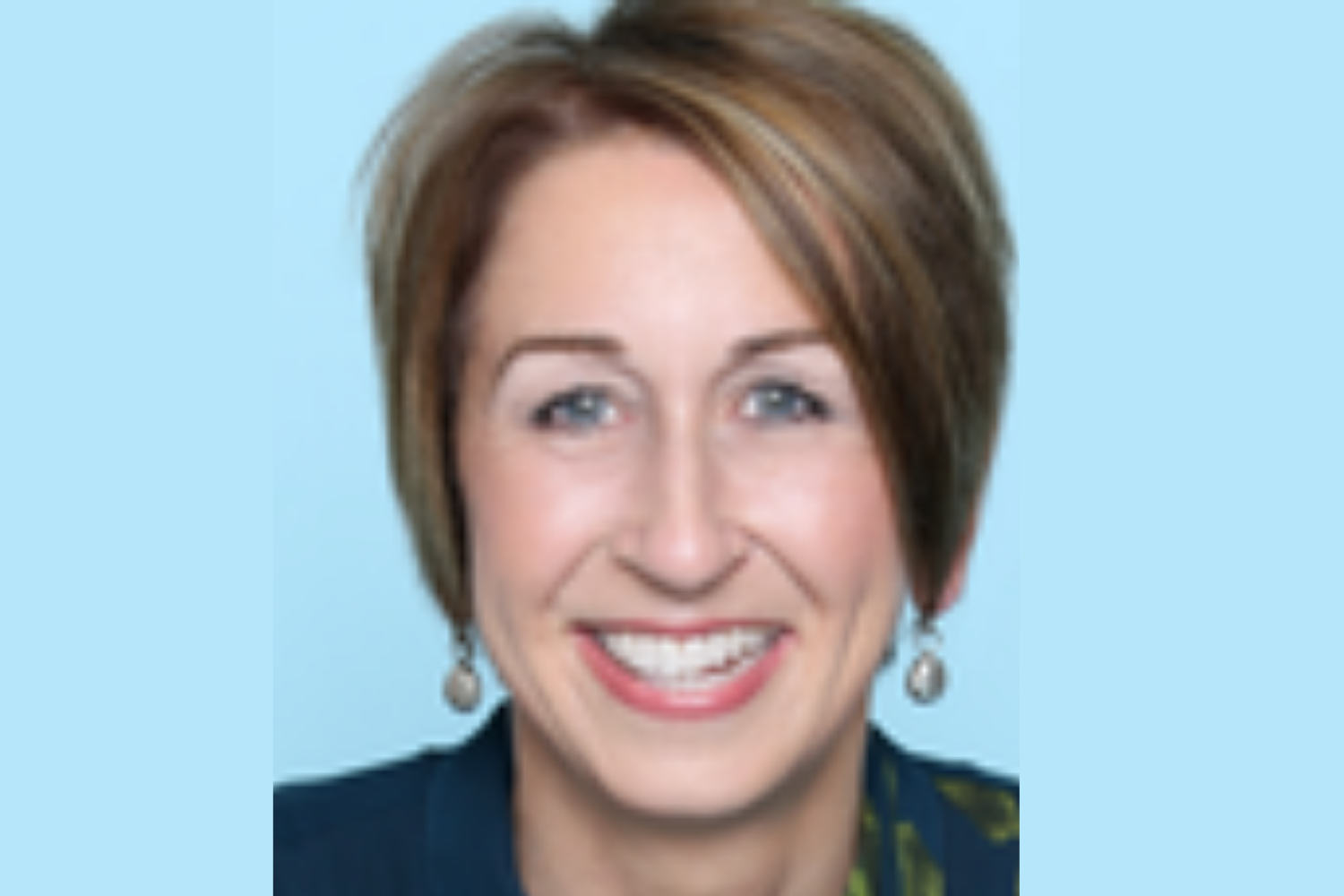 Smiling woman with short, layered brown hair, wearing earrings and a dark top, against a light blue background.