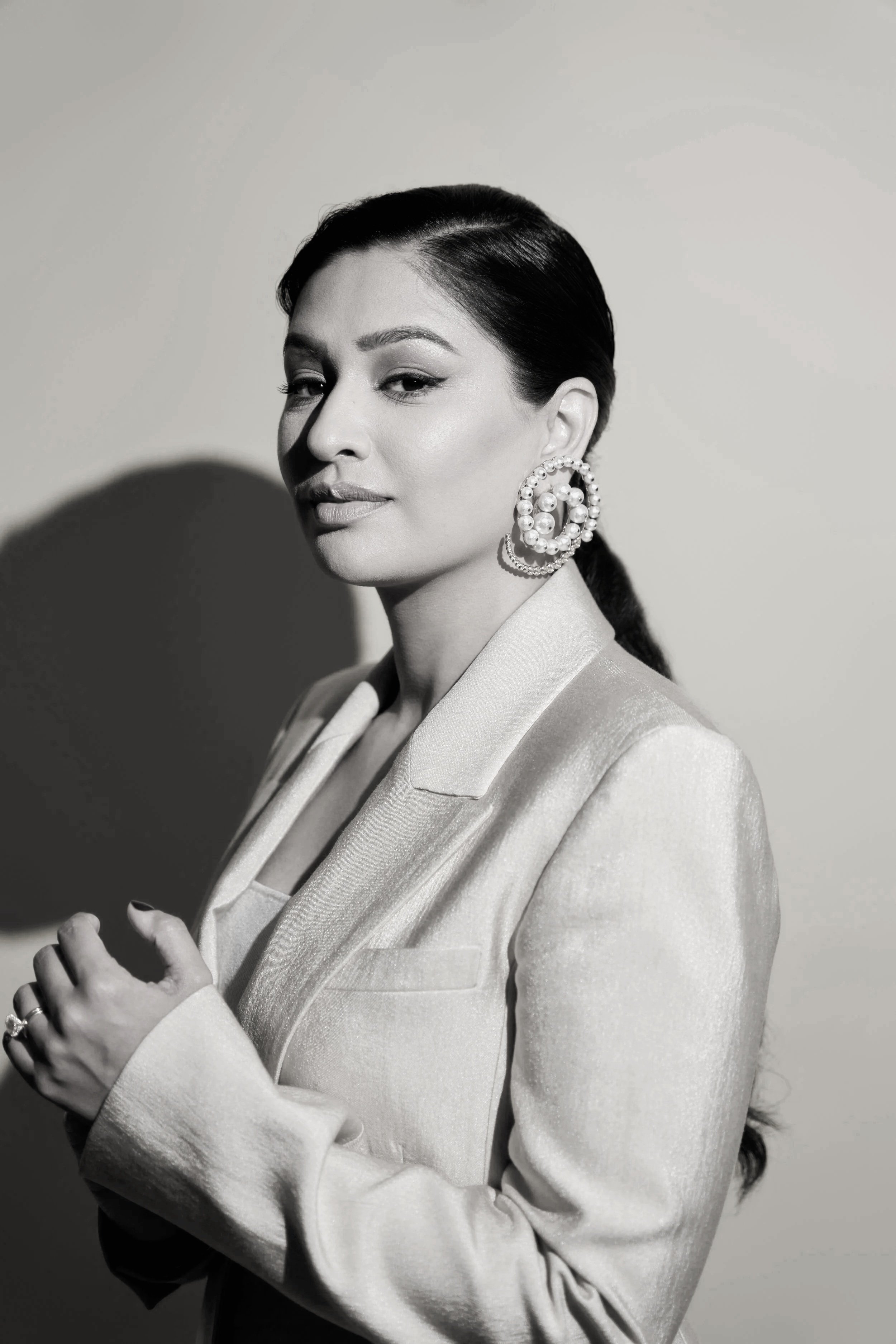 A black-and-white portrait of a woman with dark, sleek hair, wearing a light-colored blazer, large pearl earrings, and a confident expression.