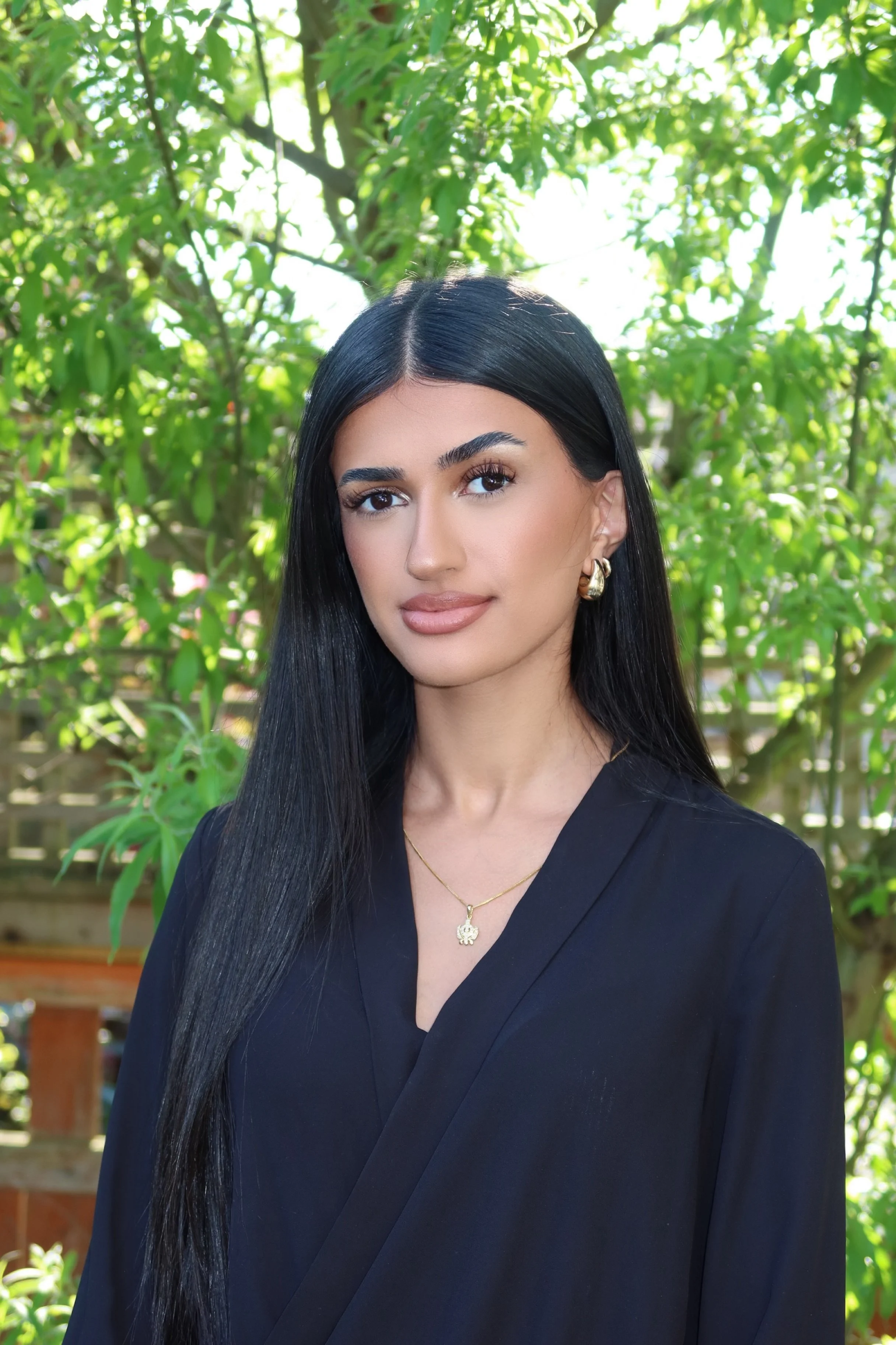 A woman with long black hair wearing gold hoop earrings and a gold necklace with a pendant, dressed in a black top or dress, standing outdoors in front of green foliage.