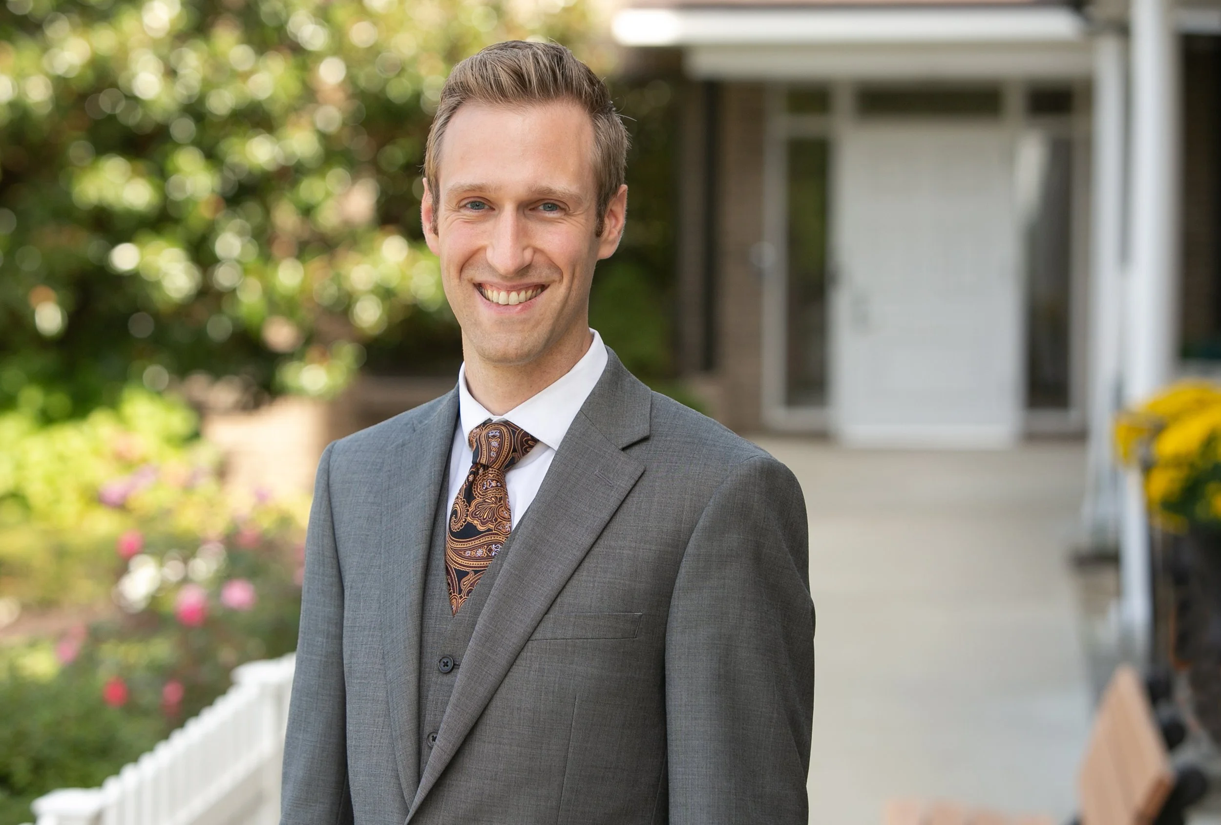 A smiling man dressed in a gray suit, white shirt, and patterned tie standing outside in front of a house with a porch and garden.