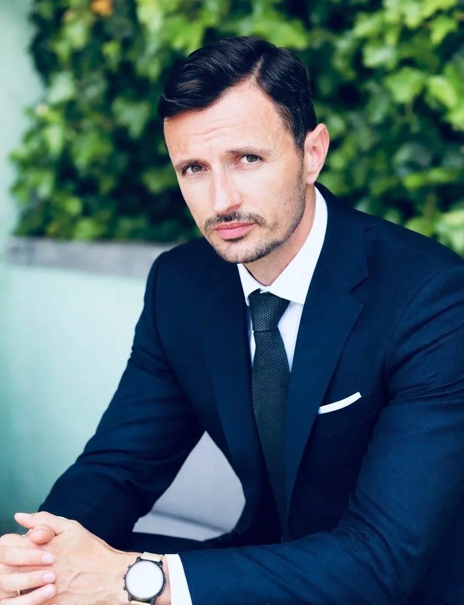 Geschäftsmann in dunklem Anzug sitzt an einem Tisch im Freien, blickt ernst in die Kamera, mit grünem Hintergrund.