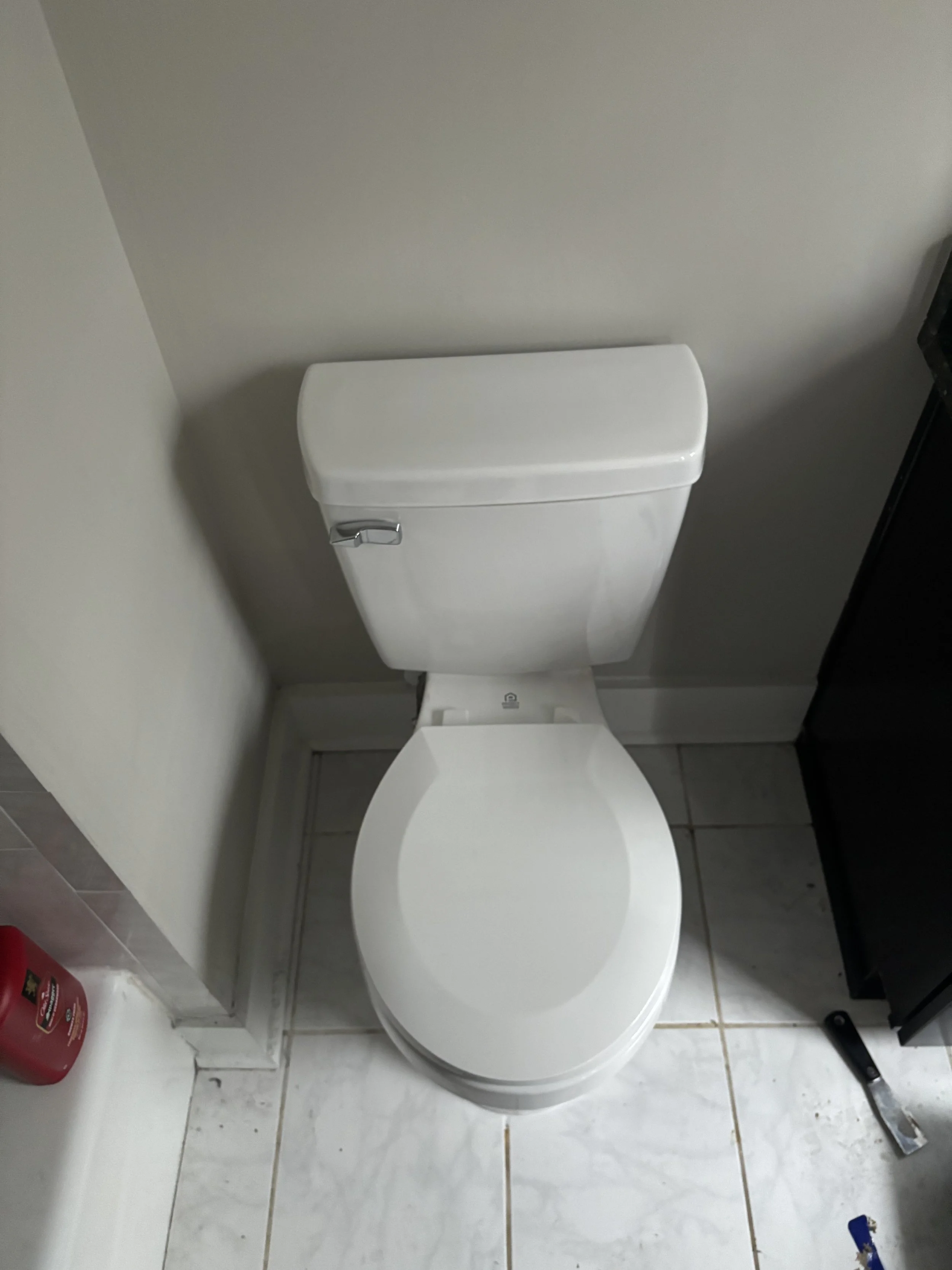 White toilet in a bathroom with tiled floor, a red fire extinguisher nearby, and a black cabinet to the right.