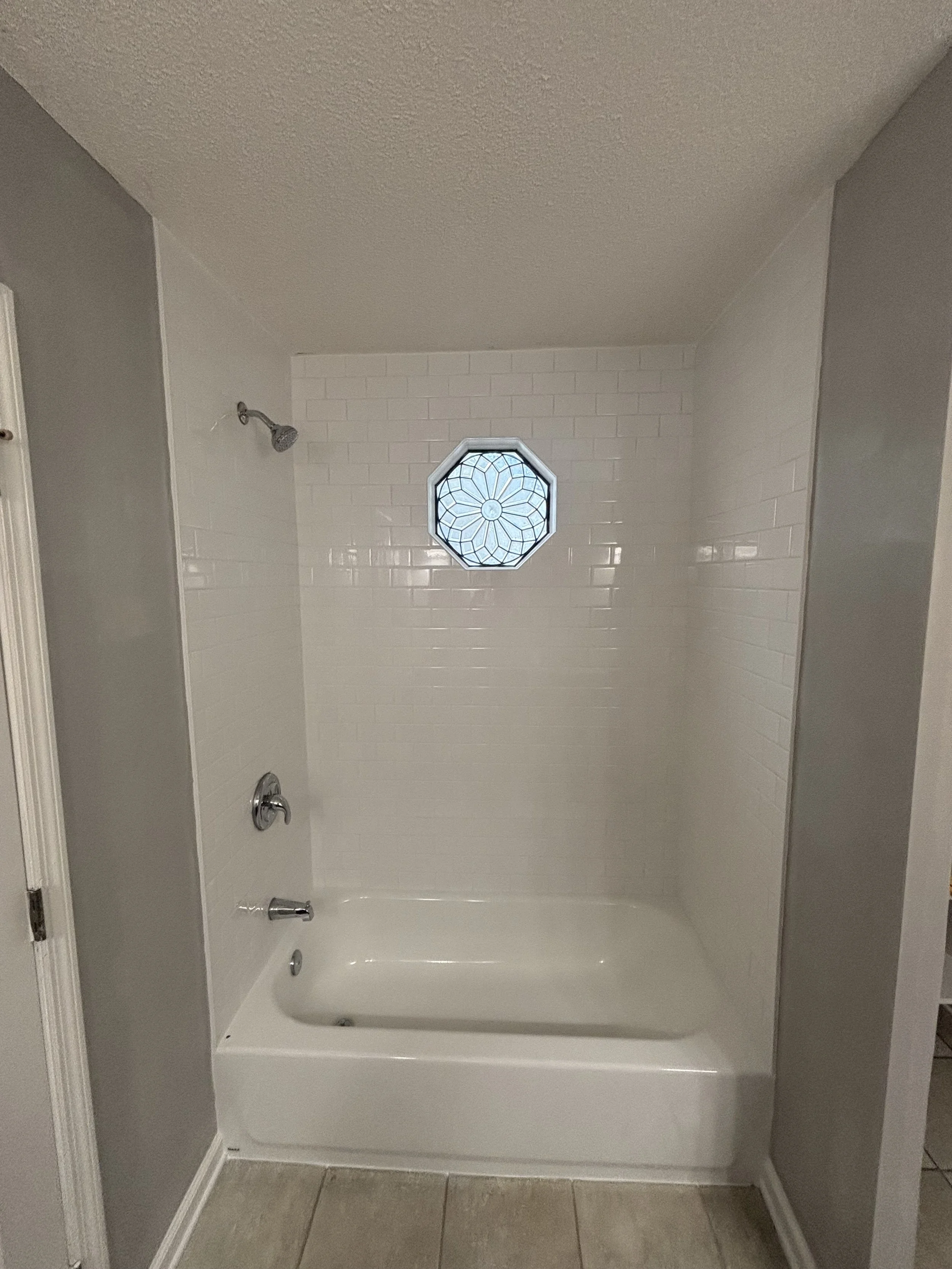 Bathroom shower with white tiled walls, a small decorative octagonal window, and a chrome showerhead.