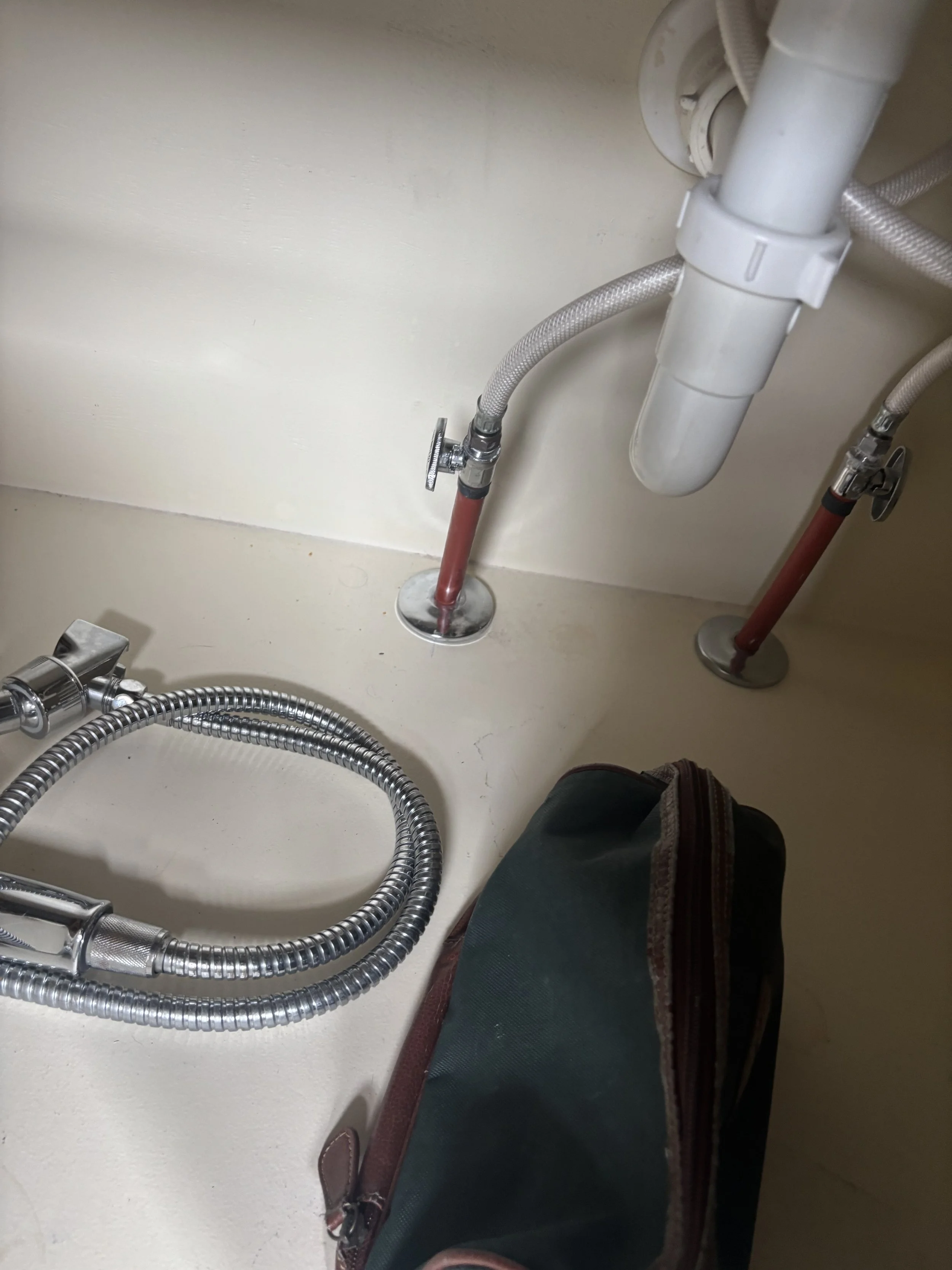 Underneath a kitchen sink showing plumbing pipes, including a white PVC pipe and flexible water supply lines, with a small bag or case nearby.