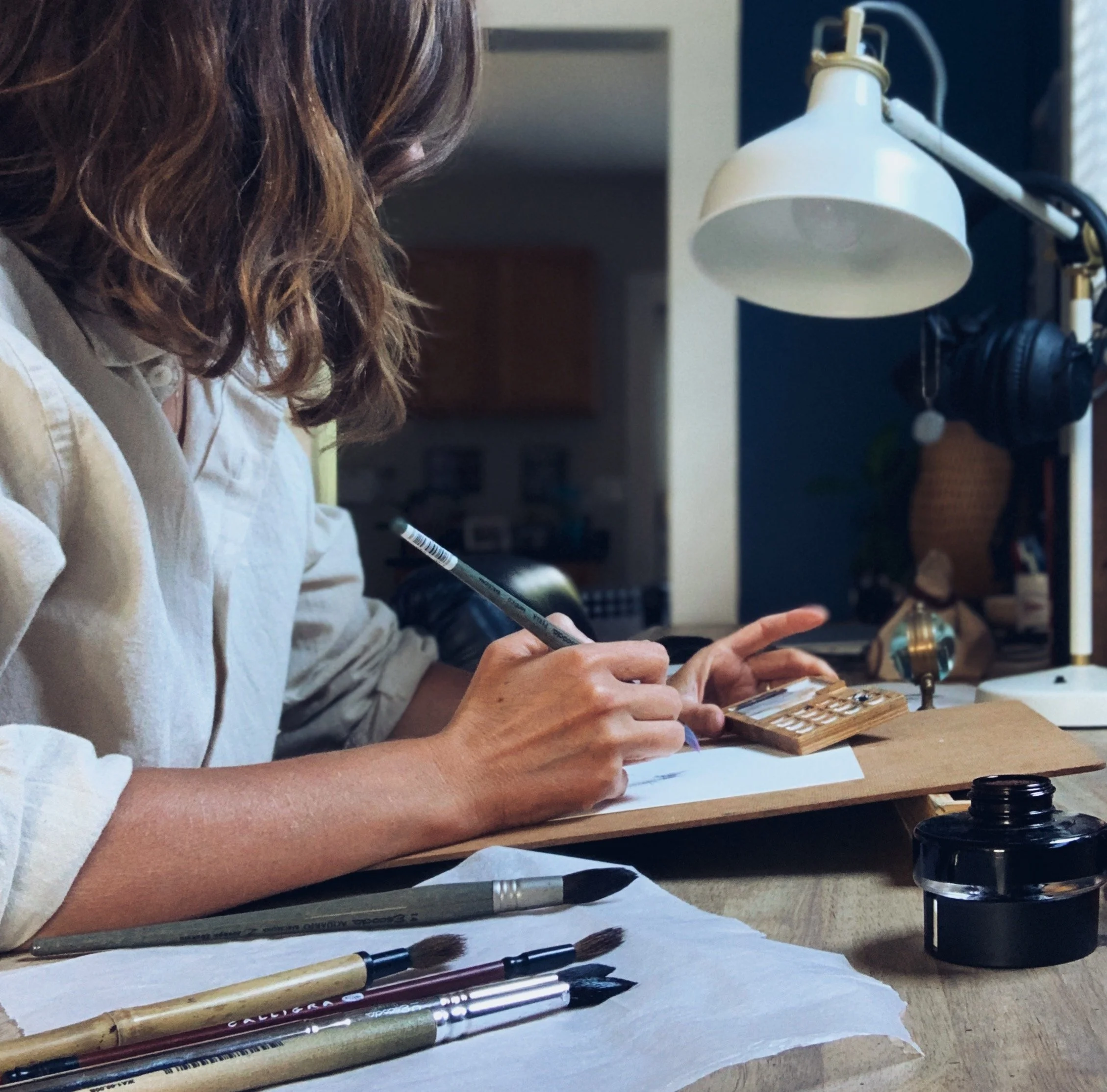 A woman with shoulder-length hair sitting at a wooden table, writing on a notepad with various paint brushes and art supplies in front of her.