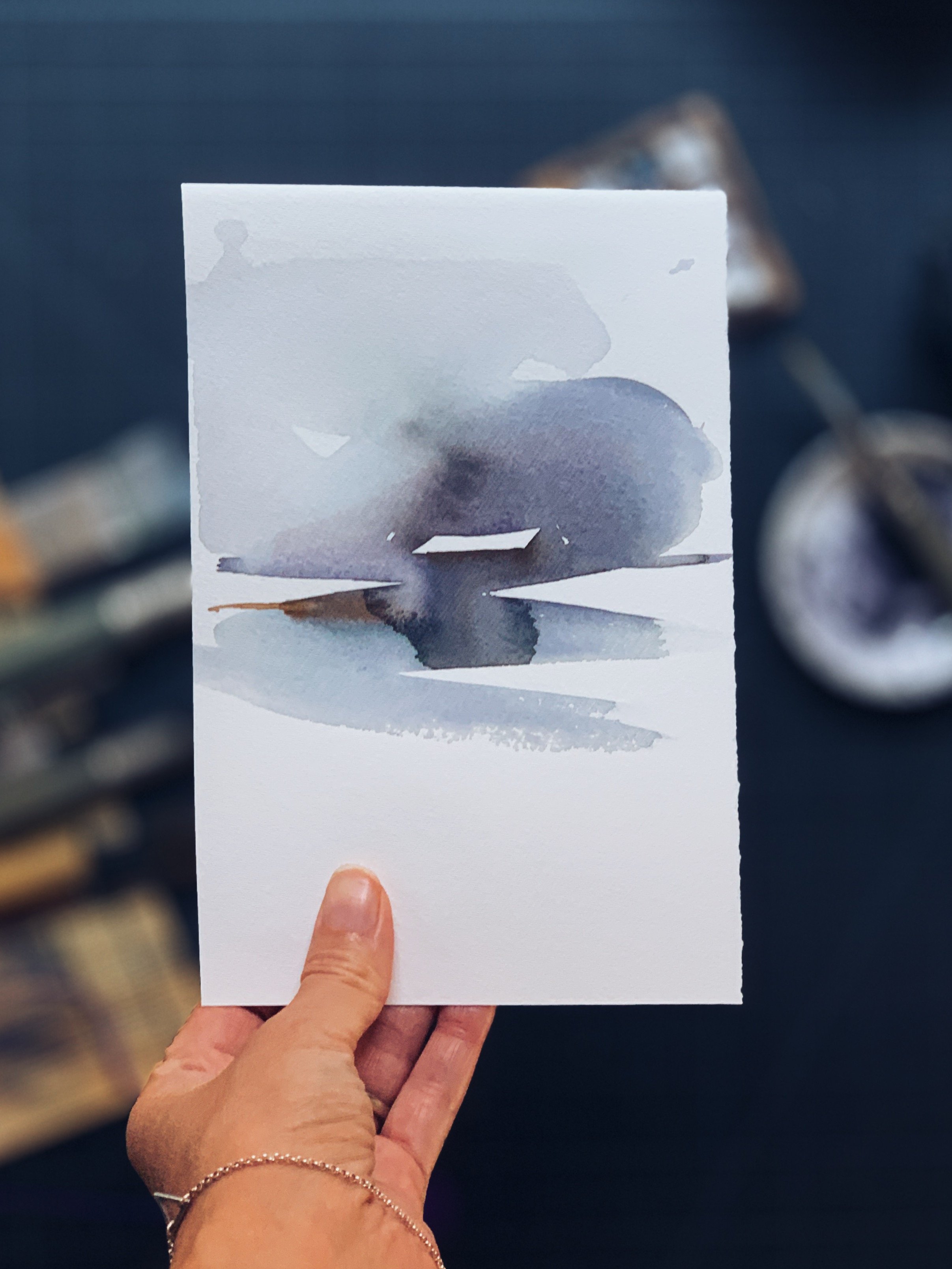 A hand holding a watercolor painting of a boat on water with a dark cloud overhead.