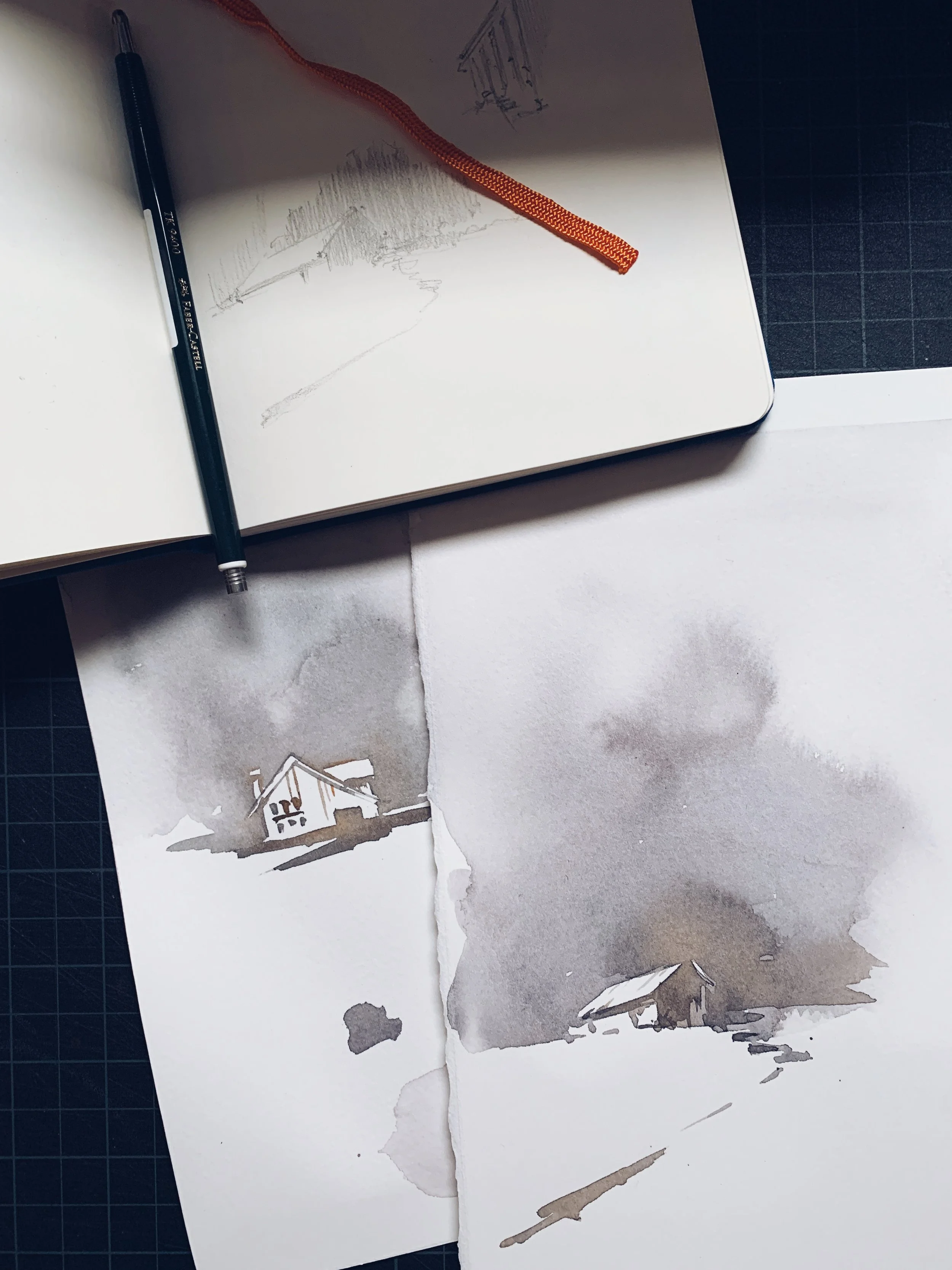 A sketchbook with a pencil and an orange ribbon bookmark on top, and a watercolor painting of a house on paper below.