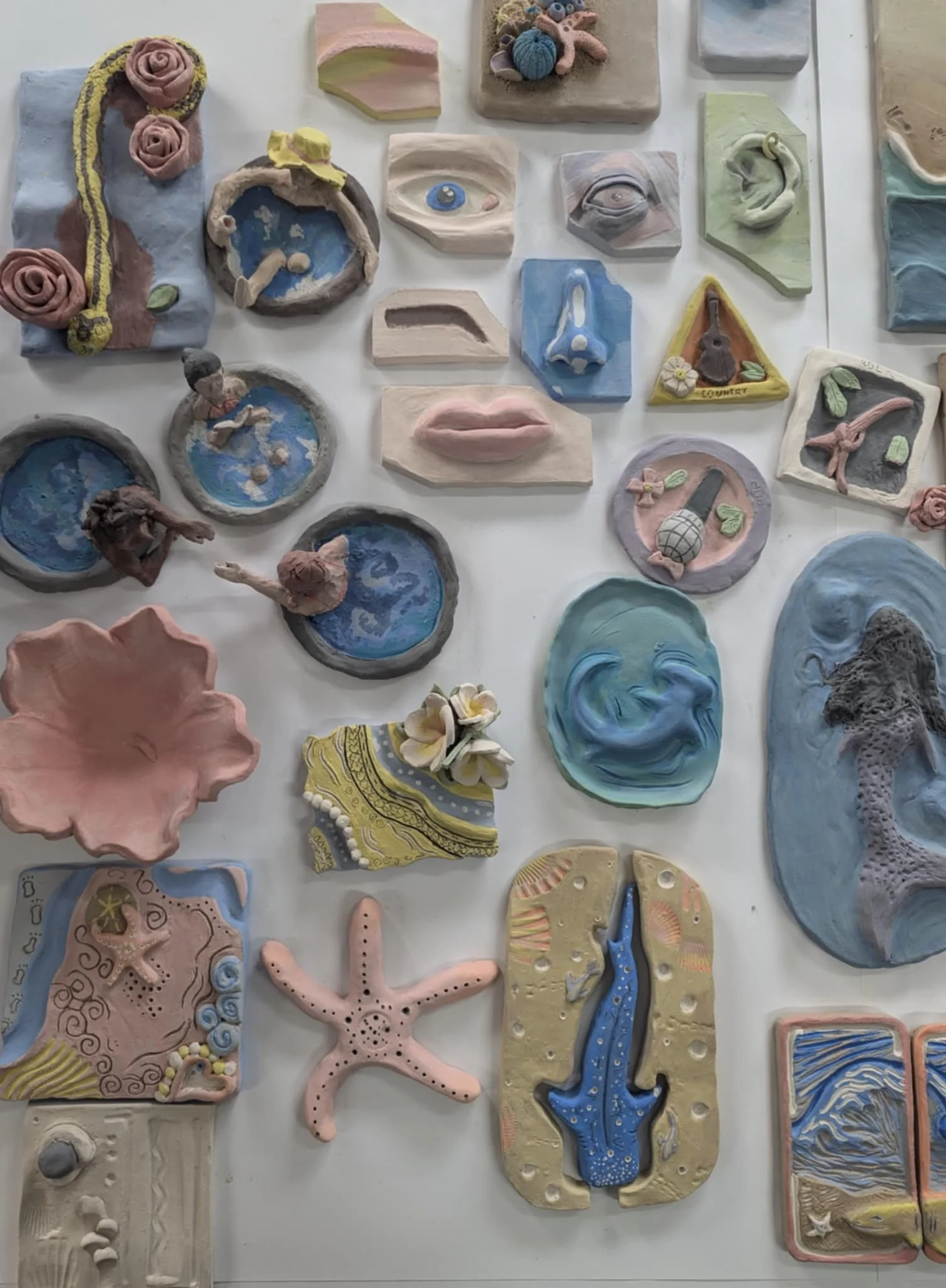 A collection of handmade clay art pieces including faces, animals, and abstract designs, with some featuring faces, fish, starfish, and ocean themes in pastel colors.