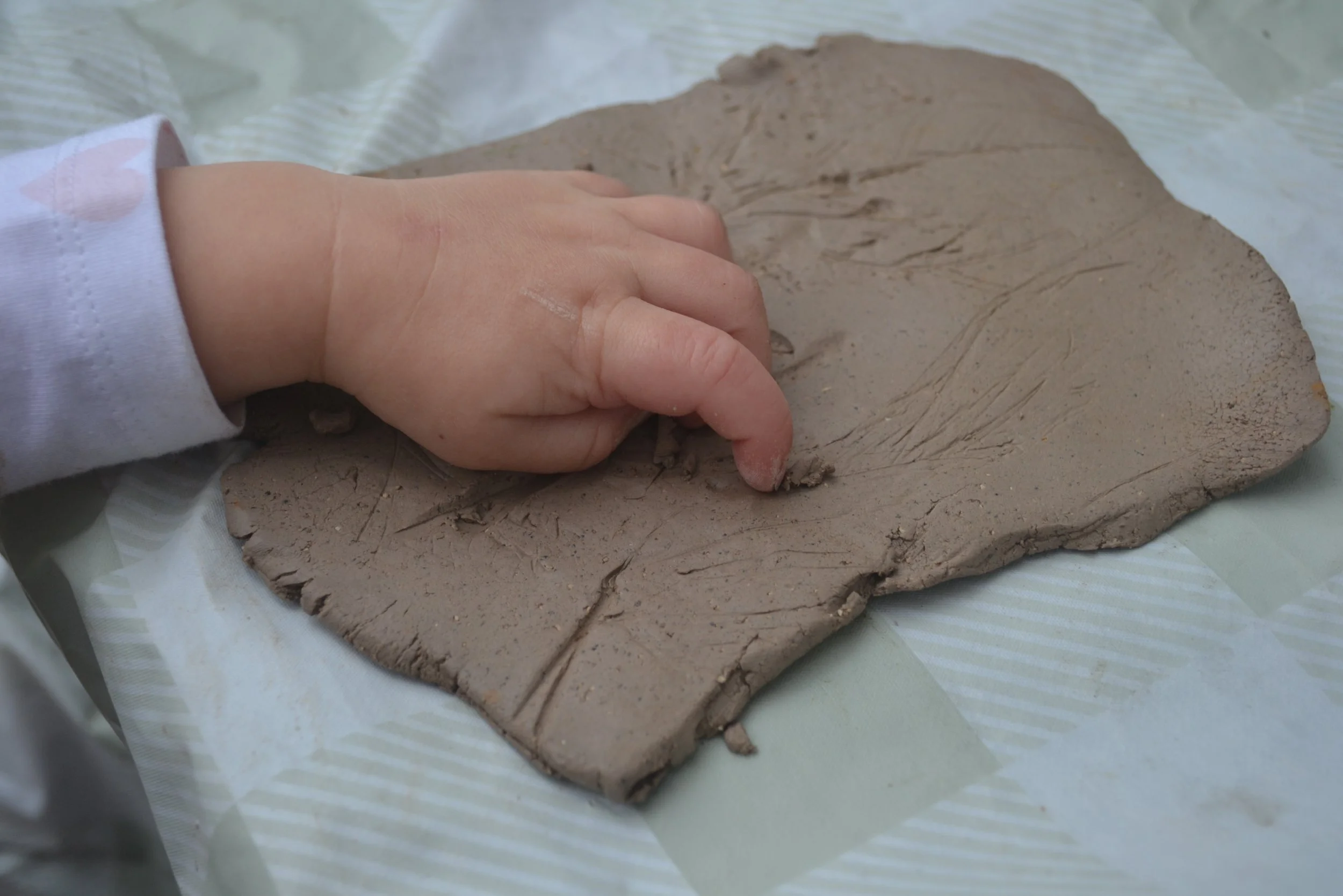 A child's hand pressing into a flat piece of modeling clay on a striped surface.