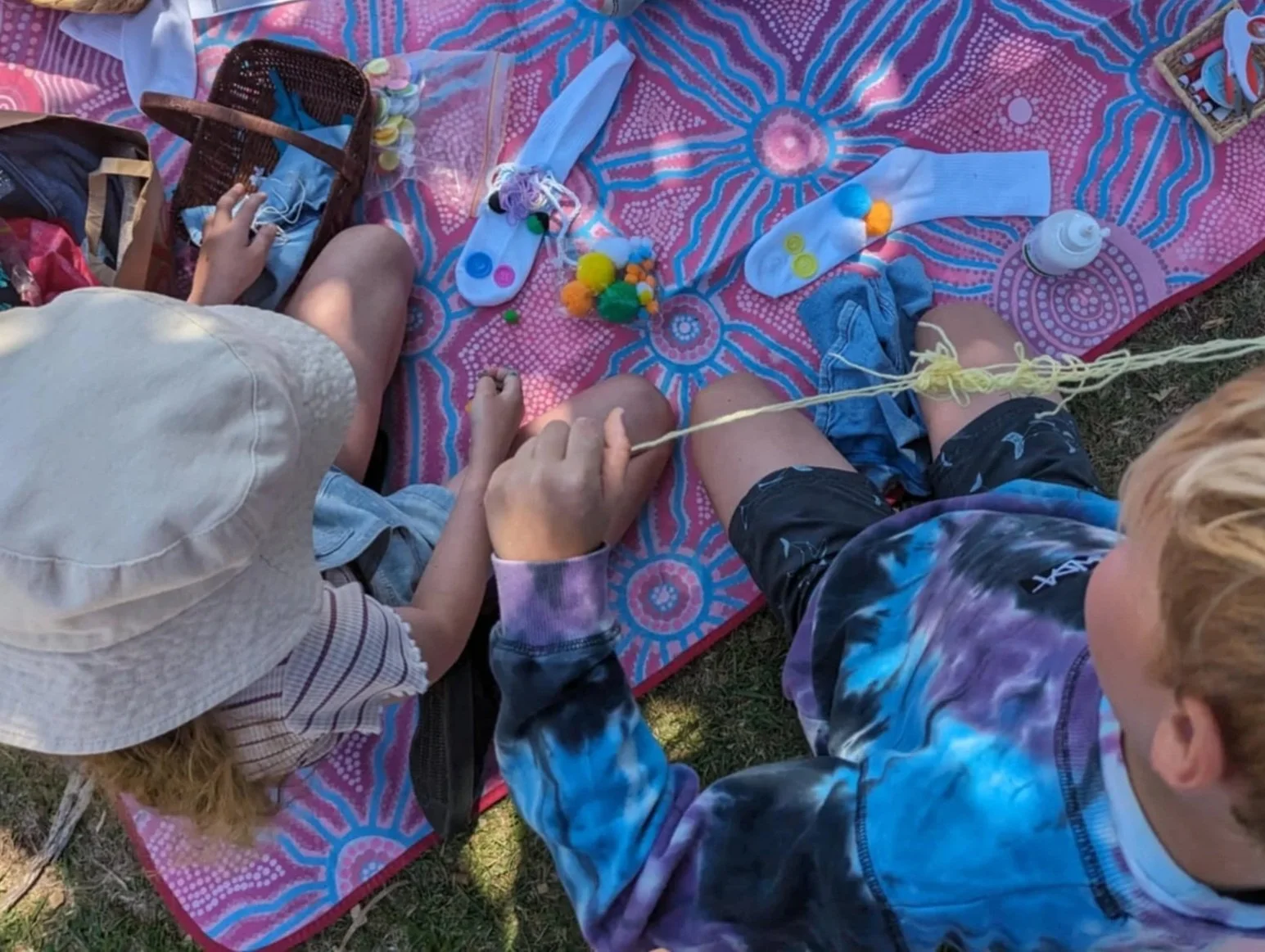 Two children sitting on a colorful blanket outdoors, engaging in arts and crafts with pom-poms, markers, and paper.