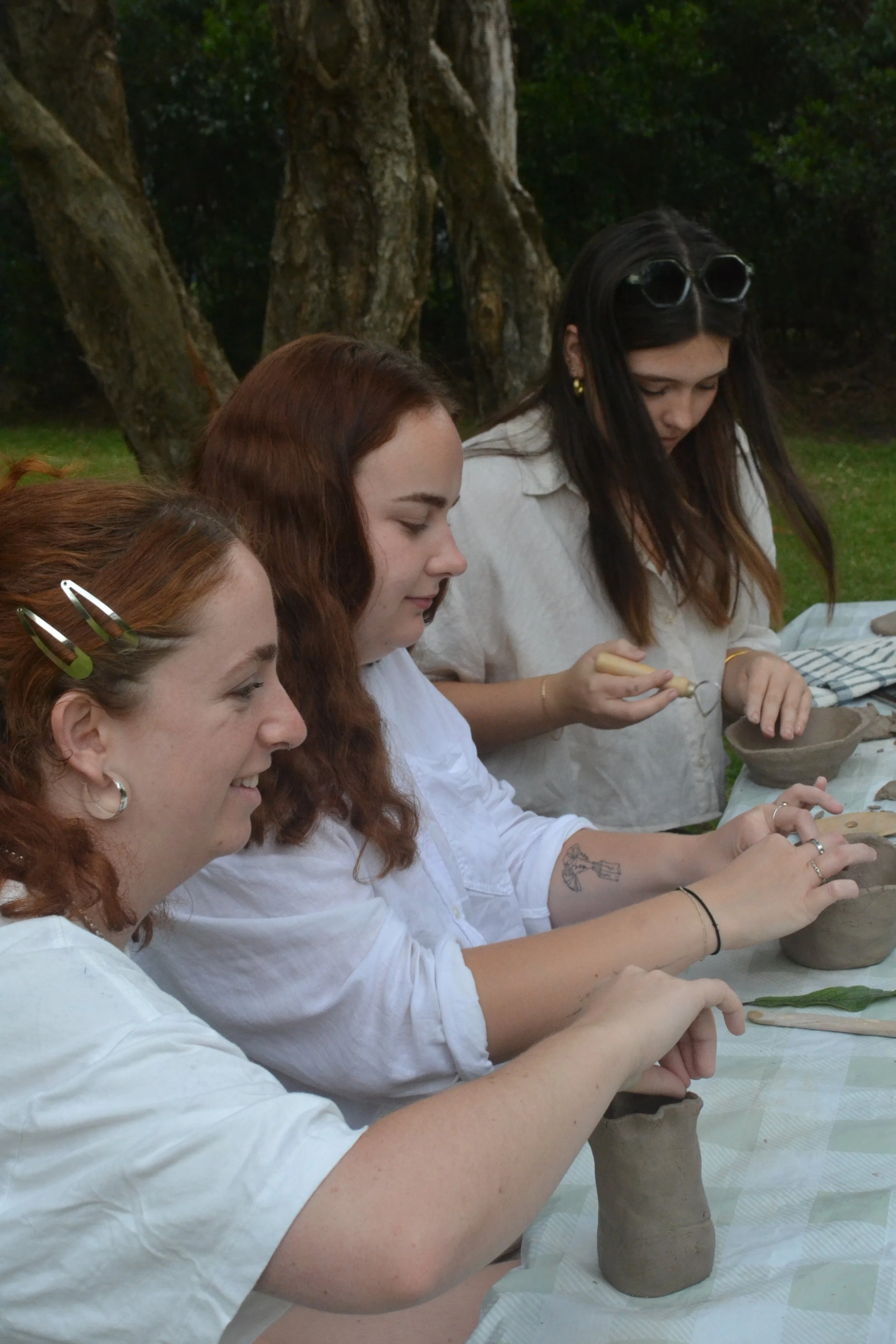 Three women sitting at a table outdoors, crafting pottery with clay and pottery tools, with trees in the background.