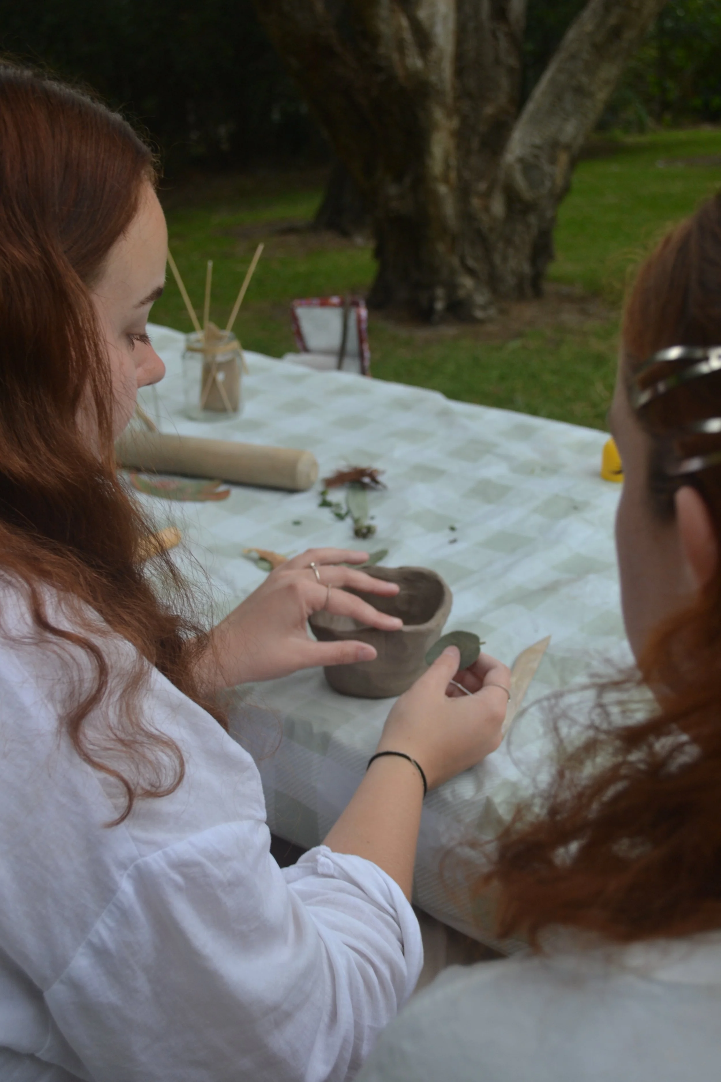 Two women with red hair creating pottery outdoors at a table covered with a white cloth, surrounded by green grass and large trees.