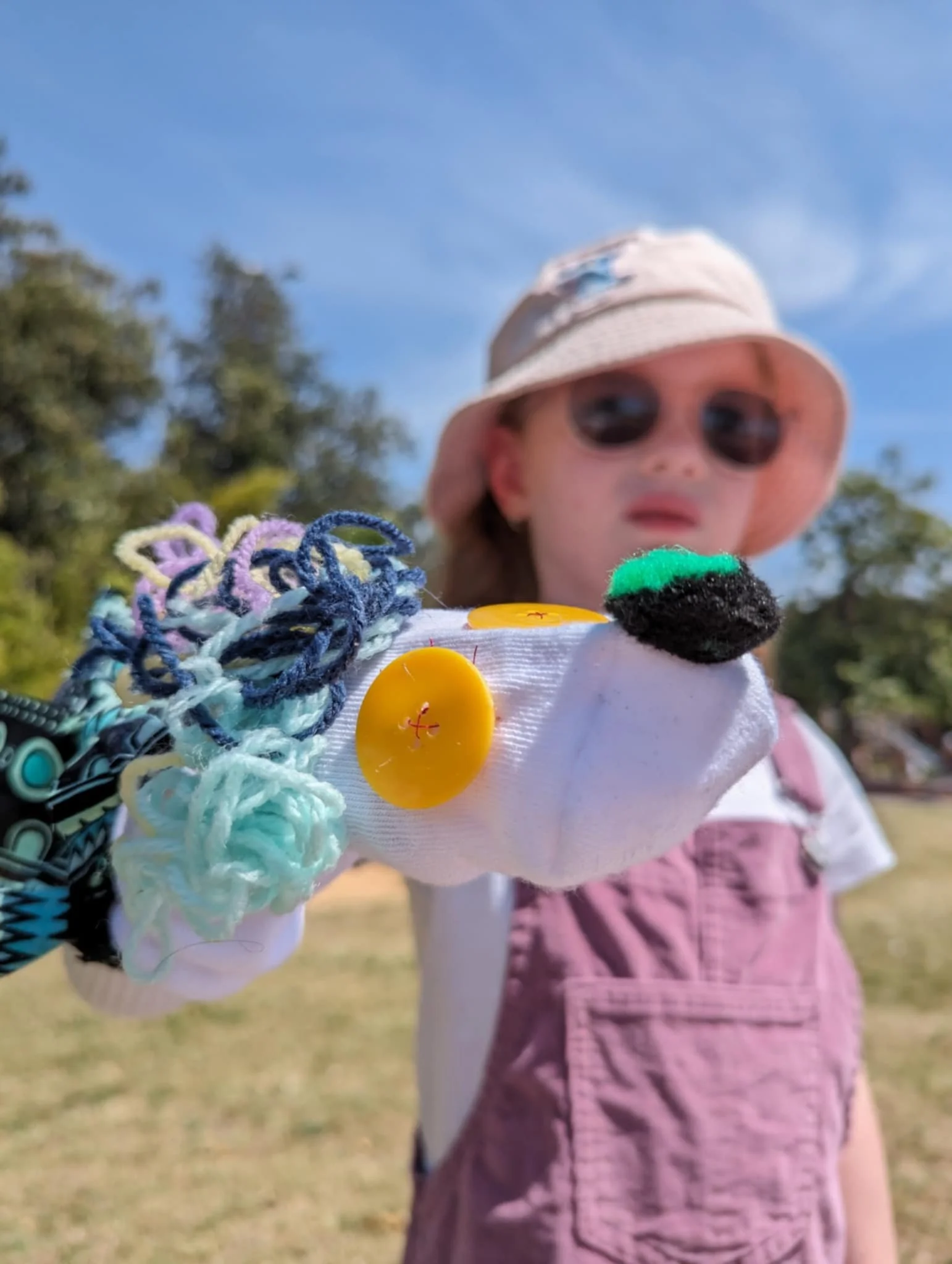 A young girl wearing a sun hat and sunglasses holding a sock puppet decorated with yarn, buttons, and a felt nose outdoors on a sunny day.