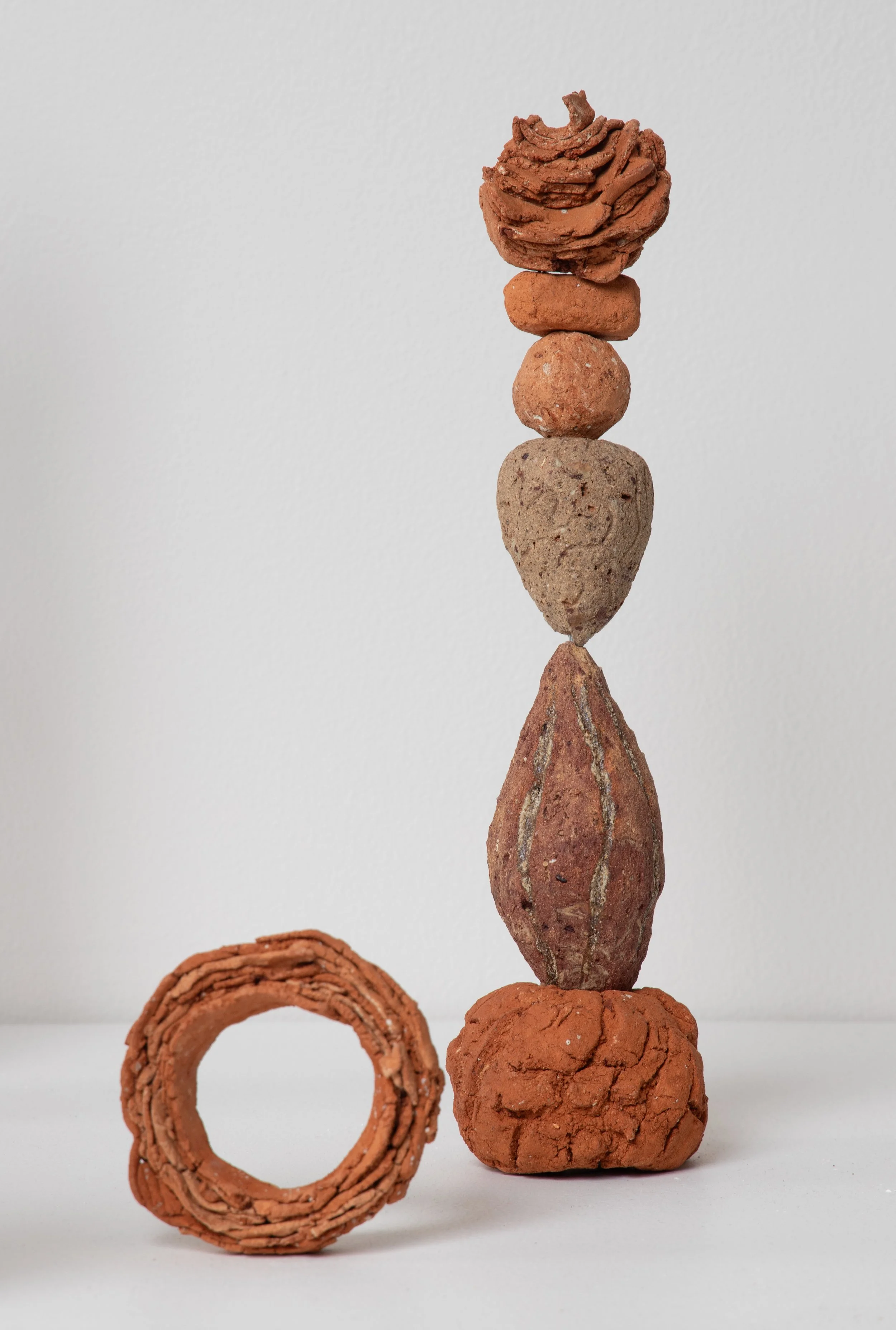 A vertical stack of wild foraged clay objects arranged on a white surface, with a circular clay structure placed on the surface in front of the stack.