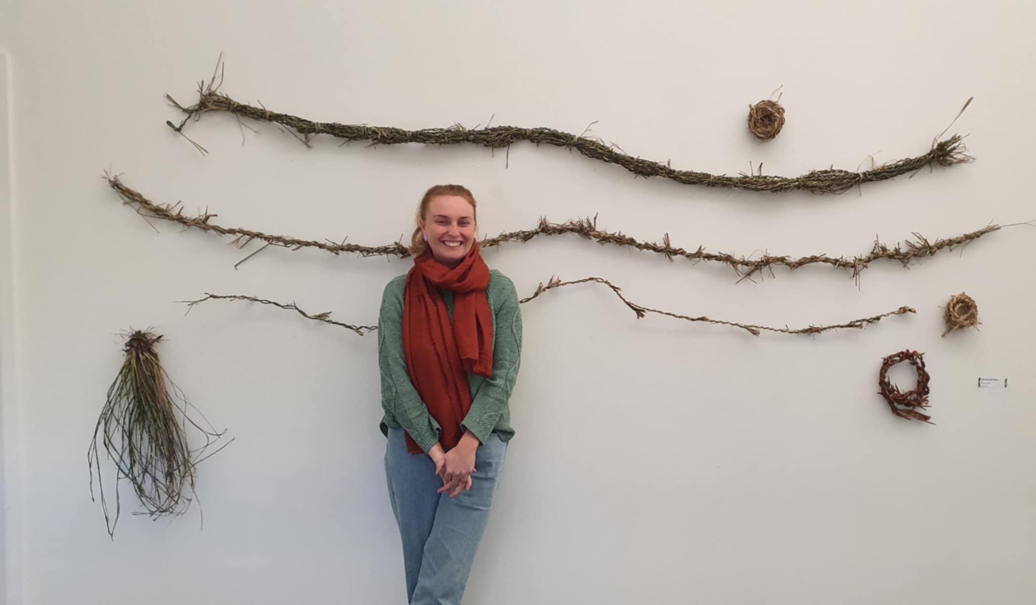 A female artist smiling and standing in front of a gallery wall decorated with long, twisted, brown natural materials arranged in wave-like patterns and three small woven wreaths.