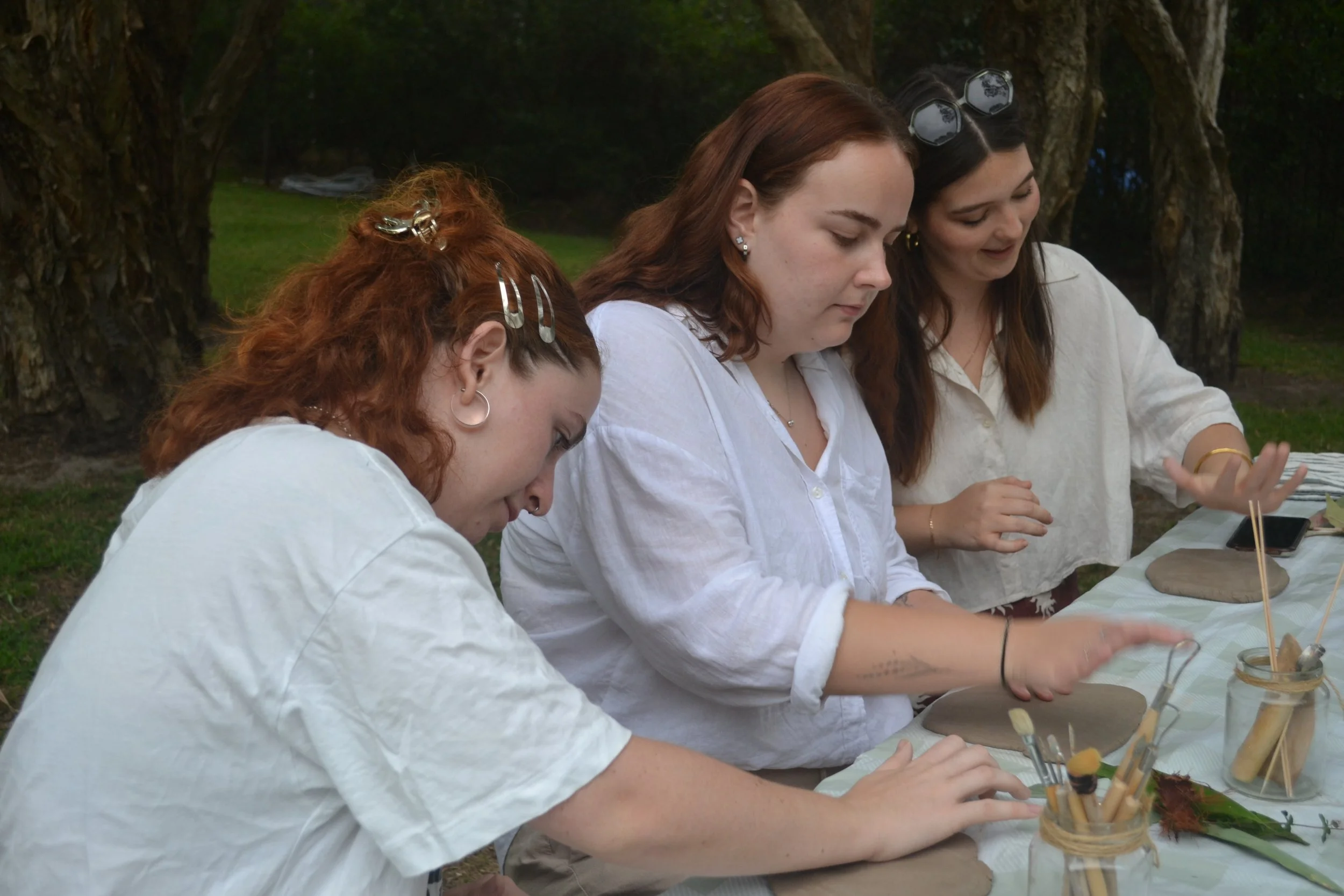 Four women working on clay projects at an outdoor table with art supplies, trees in the background.