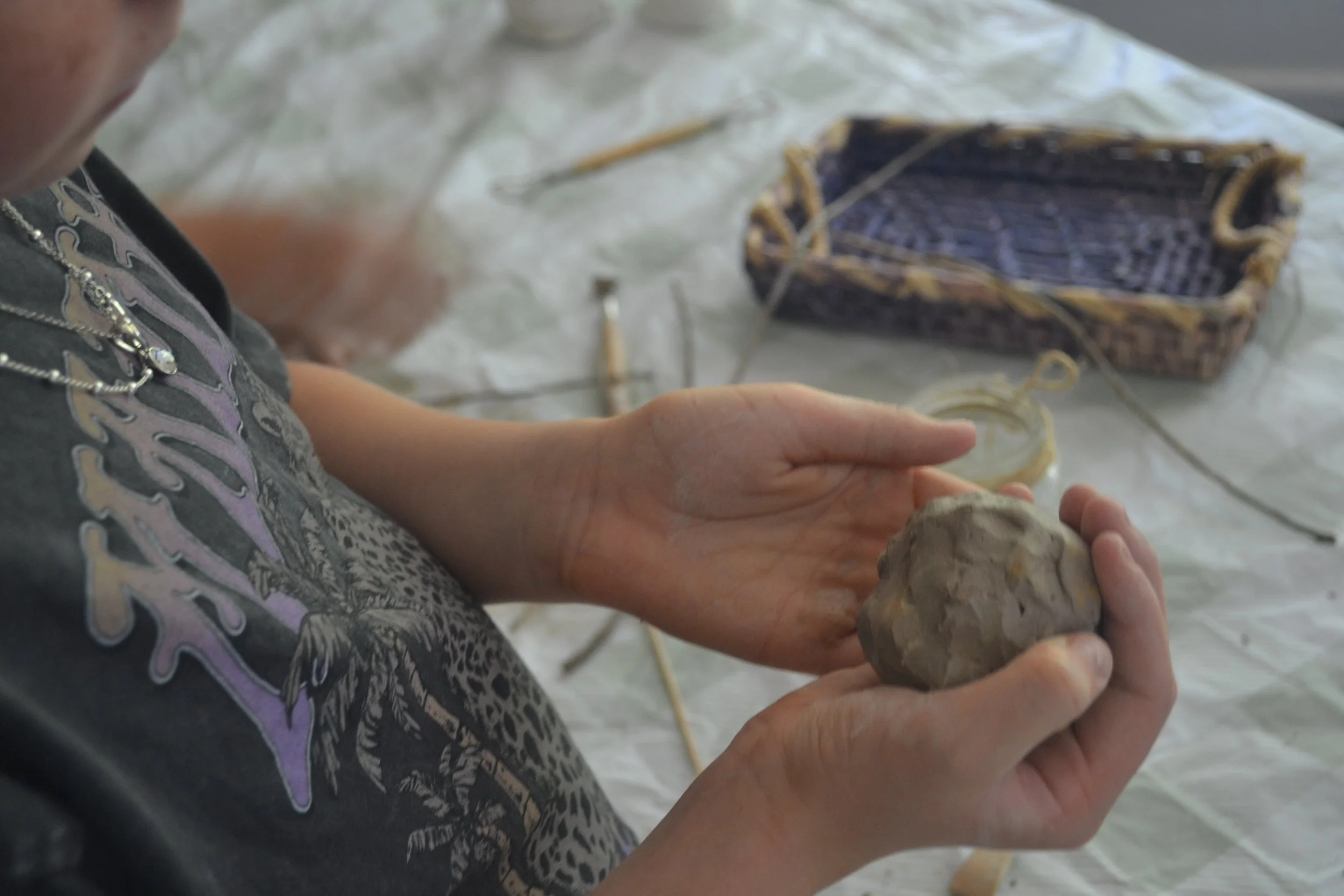 Person holding a ball of clay in their hands, with a woven basket and craft supplies on a white surface nearby.