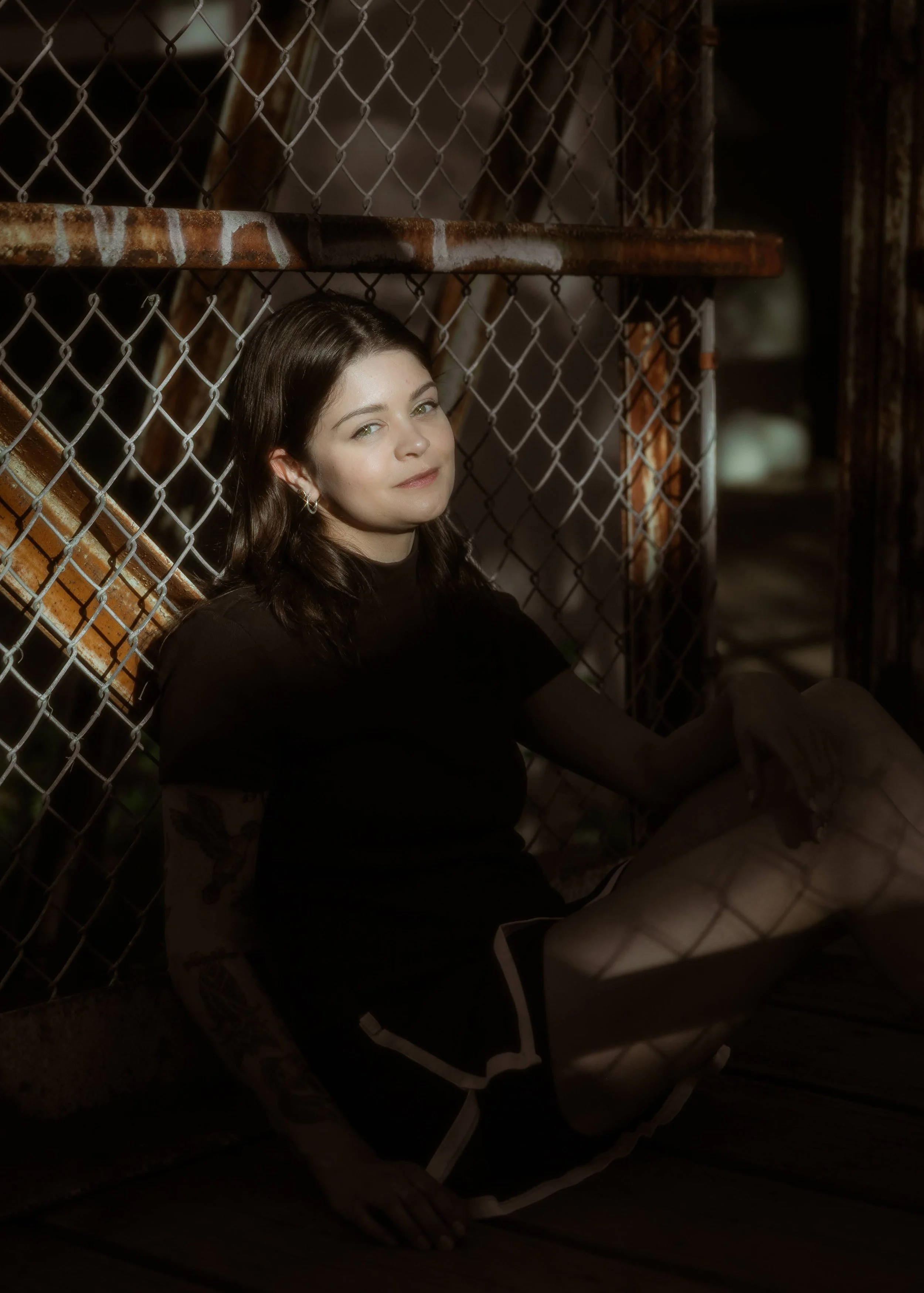 A young woman with dark hair and tattoos on her arm, sitting on the ground near a chain-link fence, illuminated by sunlight, looking at the camera with a slight smile.