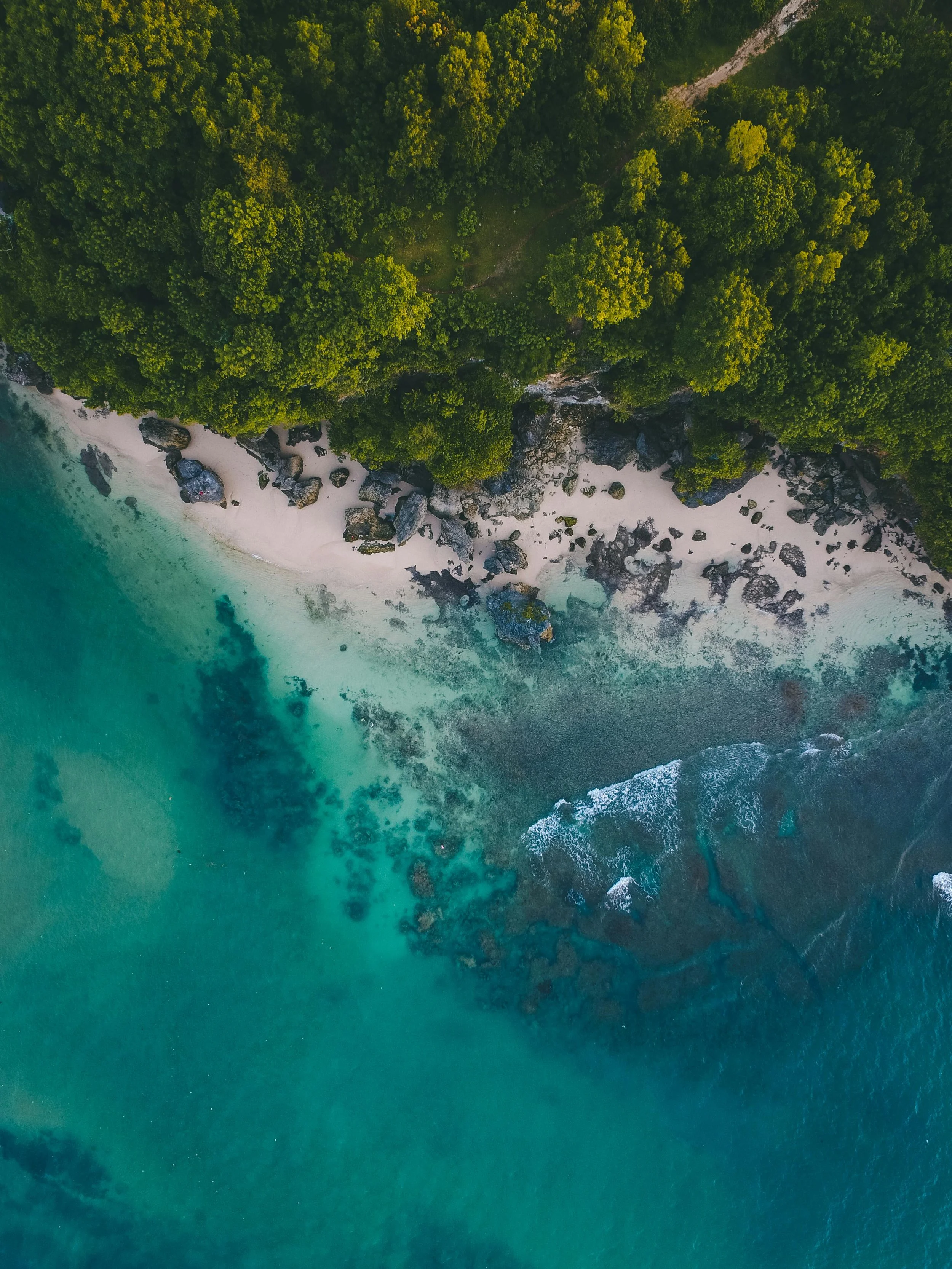 An overhead view of a beach. Rocks peek up through the white sand and teal-green waters.