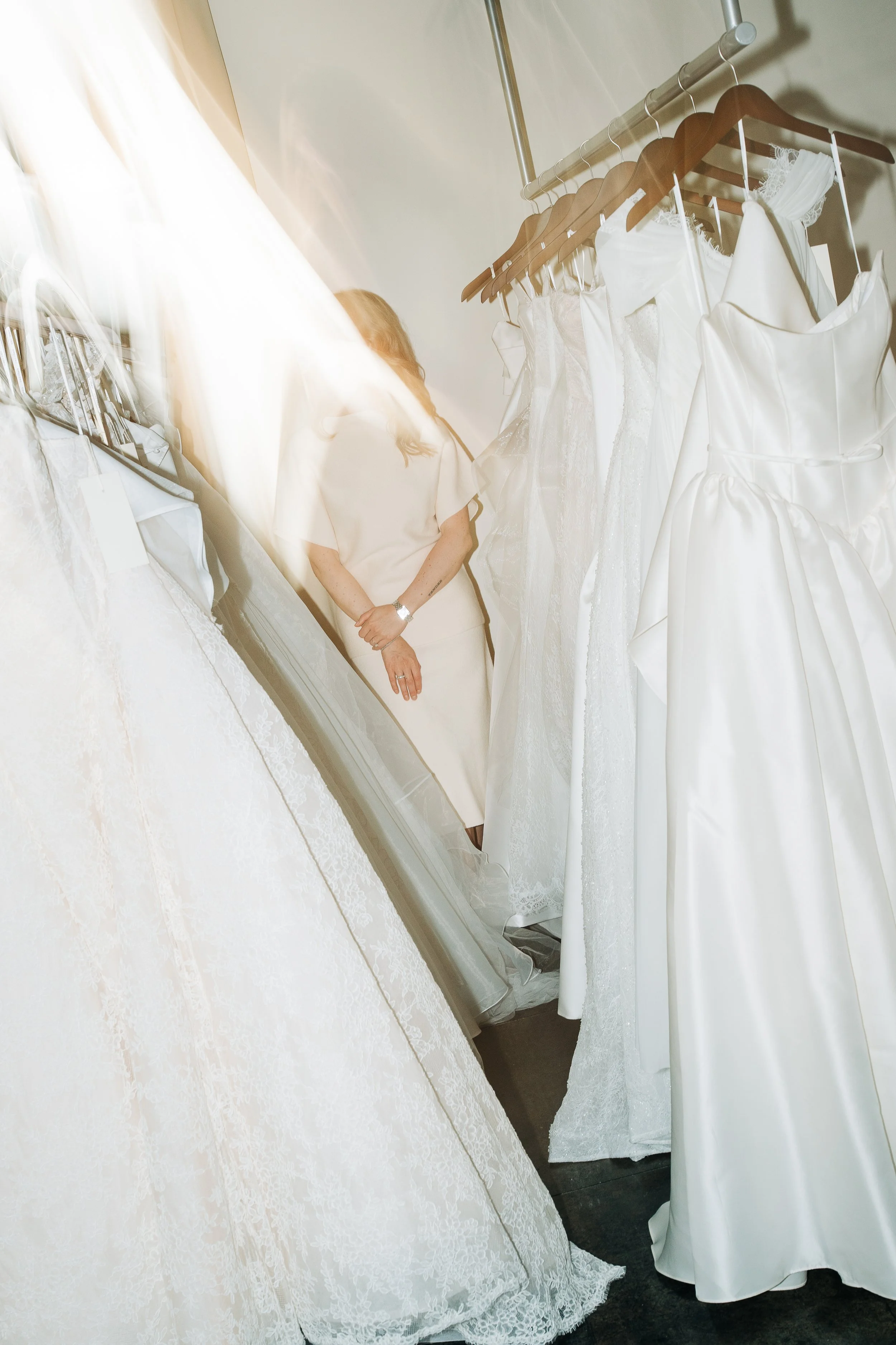 Woman trying on wedding dresses in a bridal shop, with dresses on hangers and a full-length mirror.