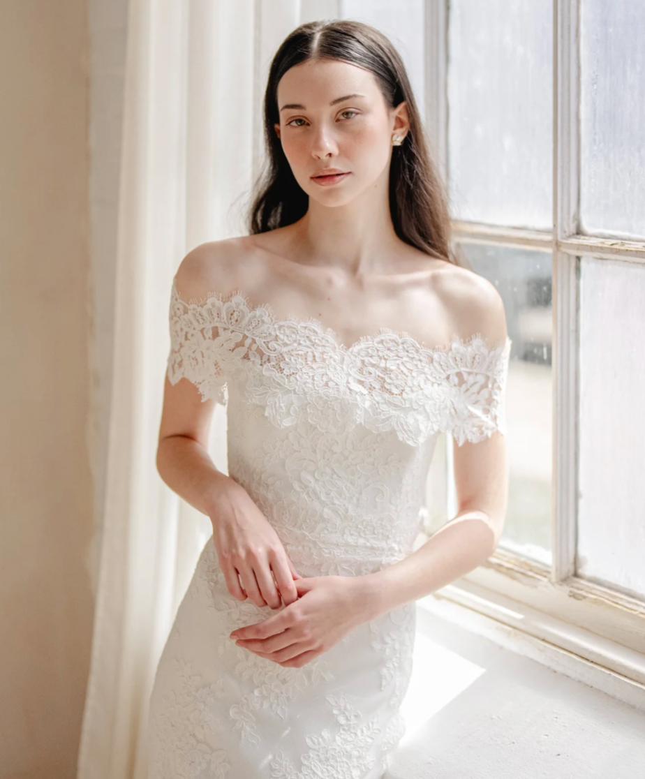 A young woman in a white lace off-shoulder dress standing by a window.