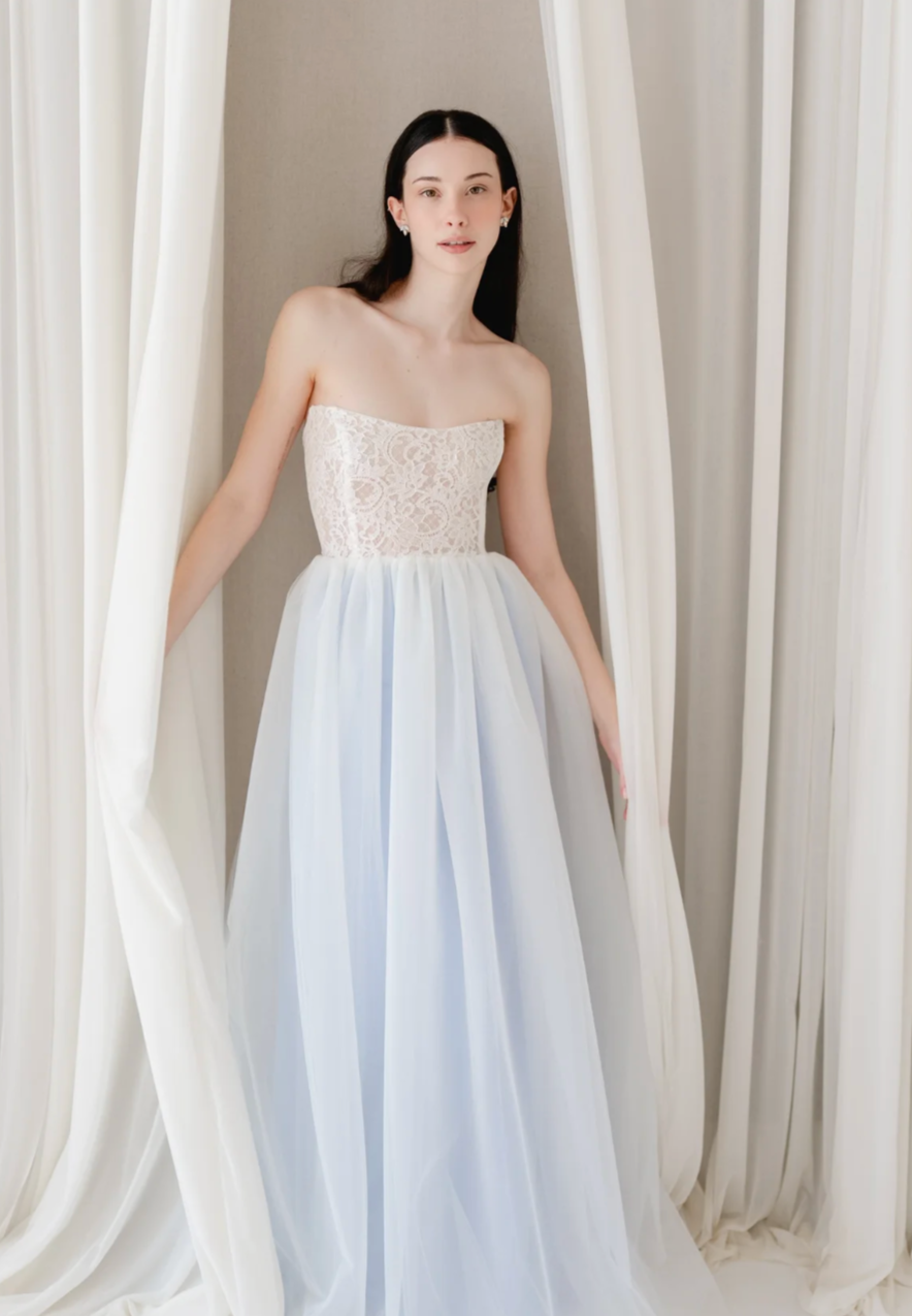 A woman standing in front of white curtains, wearing a strapless white lace top and a long, flowing white skirt.
